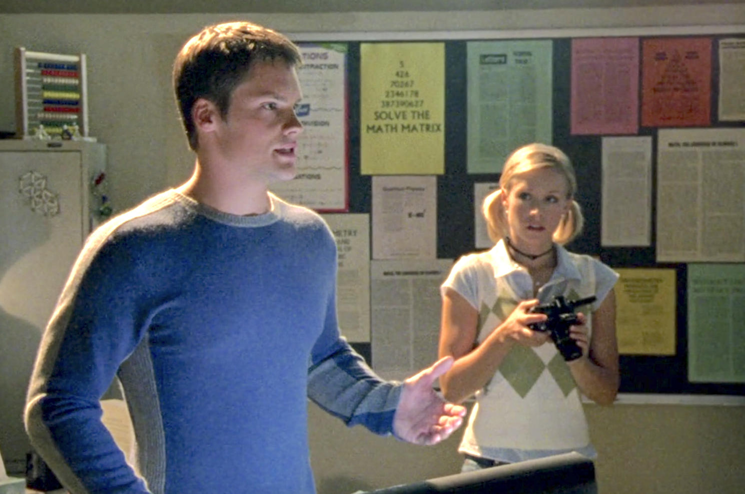 Screenshot of S1E6 of Veronica Mars. Duncan is standing at a lectern in a blue sweater giving a speech. Veronica is standing next to him with a camera watching the speech.