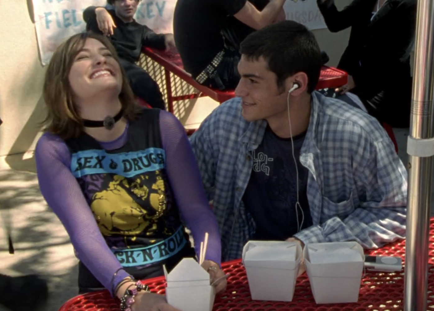 Screenshot from S1E6 of Veronica Mars. Wanda and Felix are seated at a lunch table with Chinese food takeout containers in front of them. Wanda has her head thrown back in laughter. Felix is looking at her smiling, a headphone in his ear.