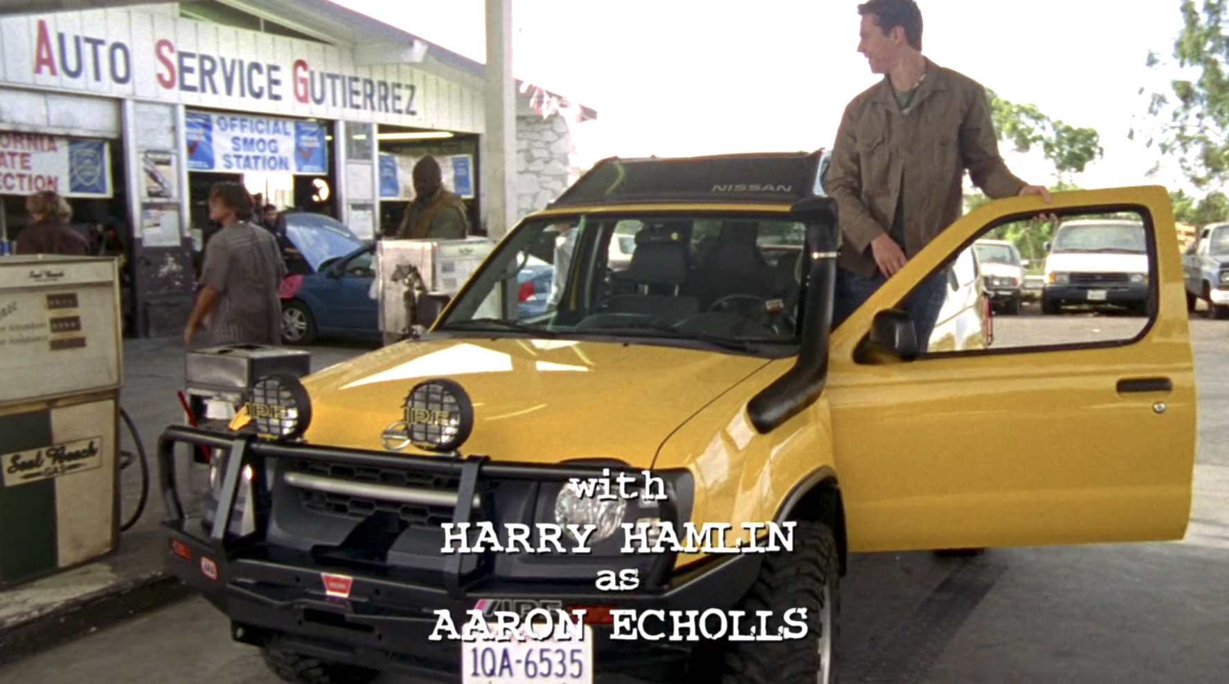 A screenshot from S1E6 of Veronica Mars. Logan has pulled his yellow SUV into a gas station. He's standing on the inside of the car, his hand on the open driver's side door, looking at his friends who are walking into the gas station.