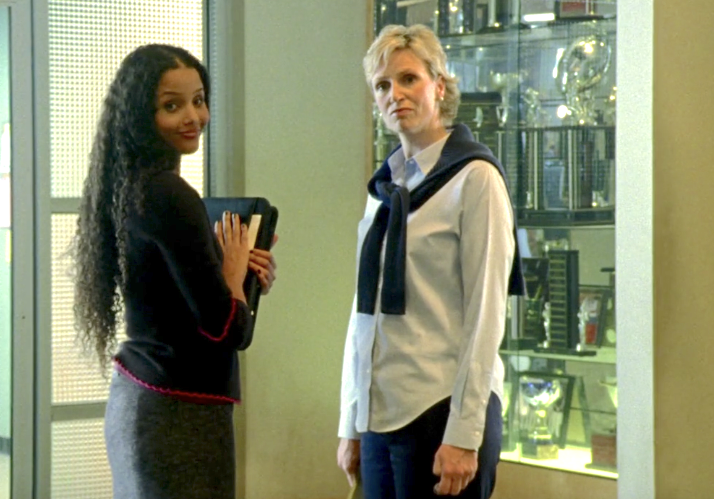 Screenshot from S1E6 of Veronica Mars. Ms. Dent and Ms. Donaldson are standing in the hallway of Neptune High. Ms. Dent has her head turned, smiling at an off-screen Veronica. Mrs. Donaldson looks perturbed.