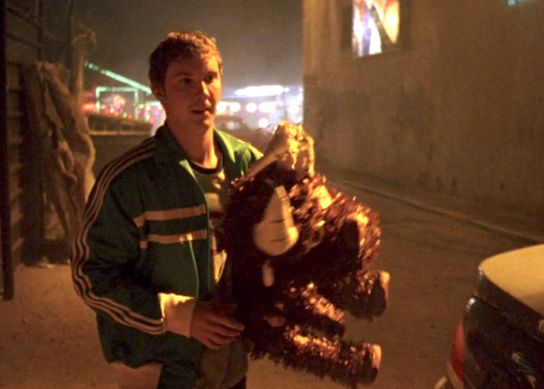 In this screenshot of S1E5, Luke, wearing a green track suit, is carrying a piñata.