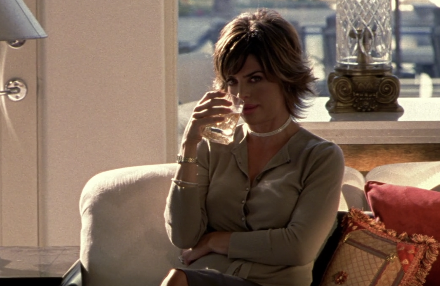 Screenshot from S1E6 of Veronica Mars. Lisa Rinna as Lynn Echolls is sitting on a white couch. Beside her are colorful pillows. She has one arm across her stomach and in her other hand she's holding a glass of brown liquor up to her mouth and staring directly into the camera.