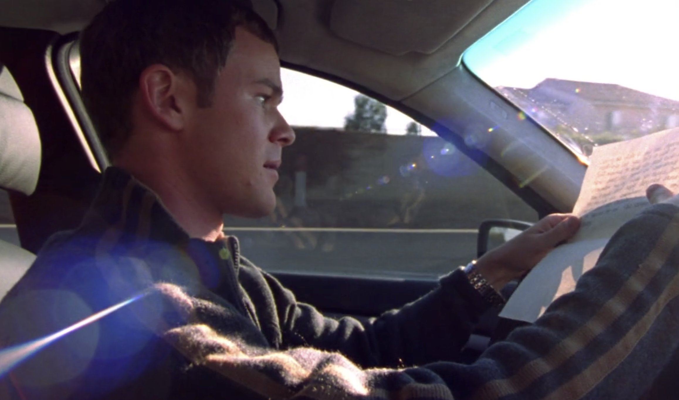 A screenshot from S1E5 of Veronica Mars. Troy is driving a car and holding a handwritten letter from Veronica, his hands leaning on the steering wheel. He looks perturbed.