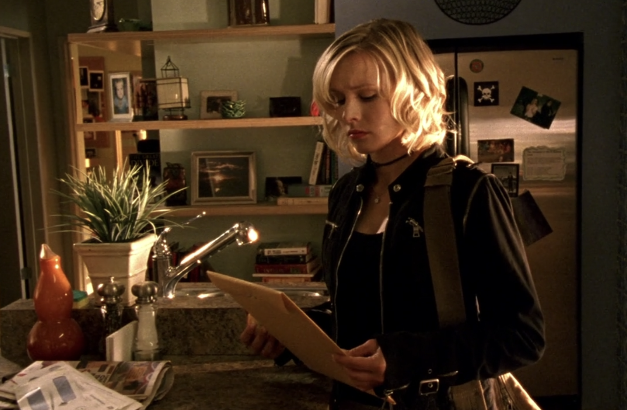 Screenshot from S1E5. Veronica is standing int he kitchen of the Mars home looking at a letter-sized yellow envelope. She looks liks she's trying to decide whether to open it.