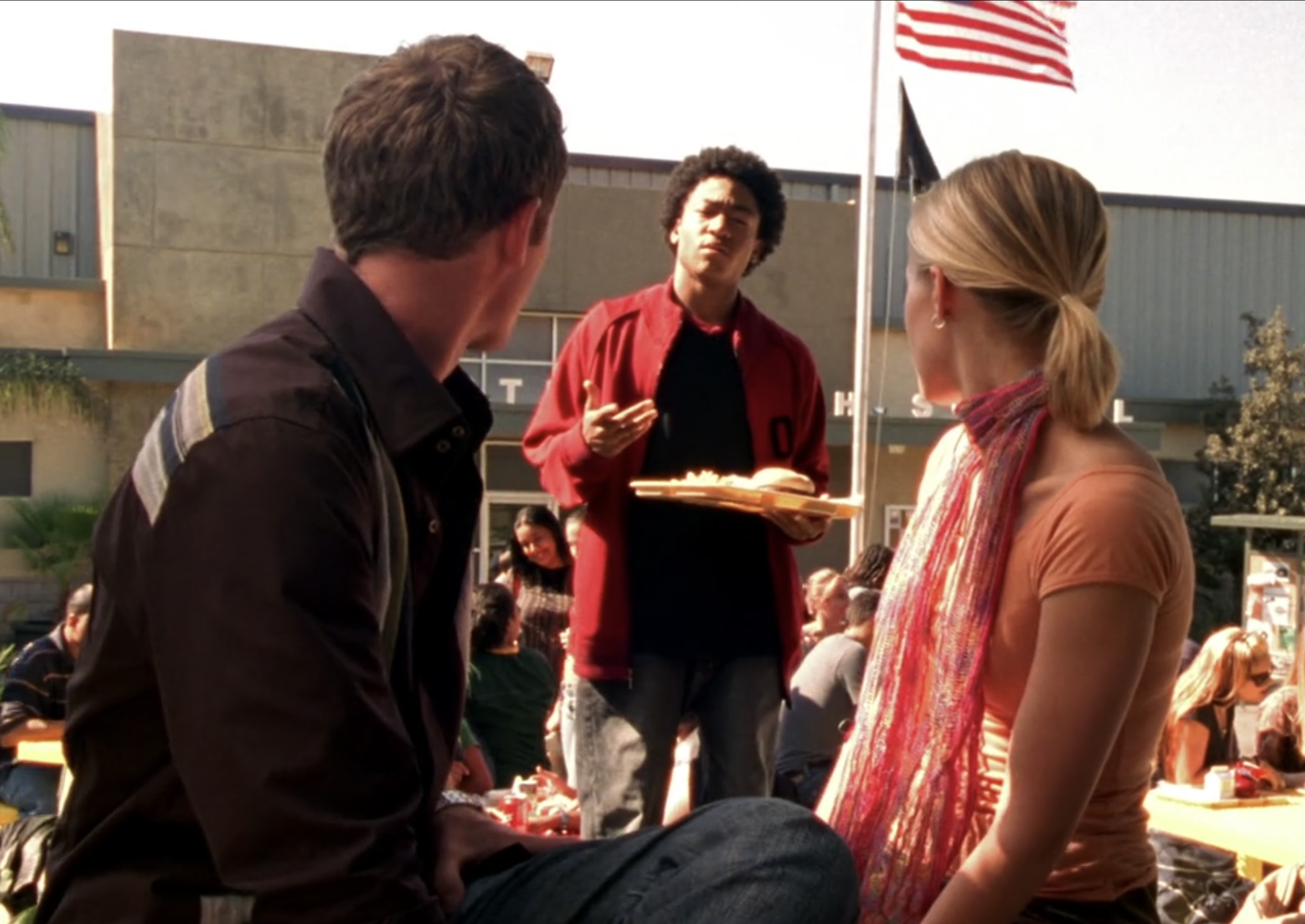 A screenshot from S1E5 of Veronica Mars. Wallace is in the background in a red jacket holding a lunch tray talking to Troy and Veronica who are in the foreground seated next to each other and looking behind them at Wallce.