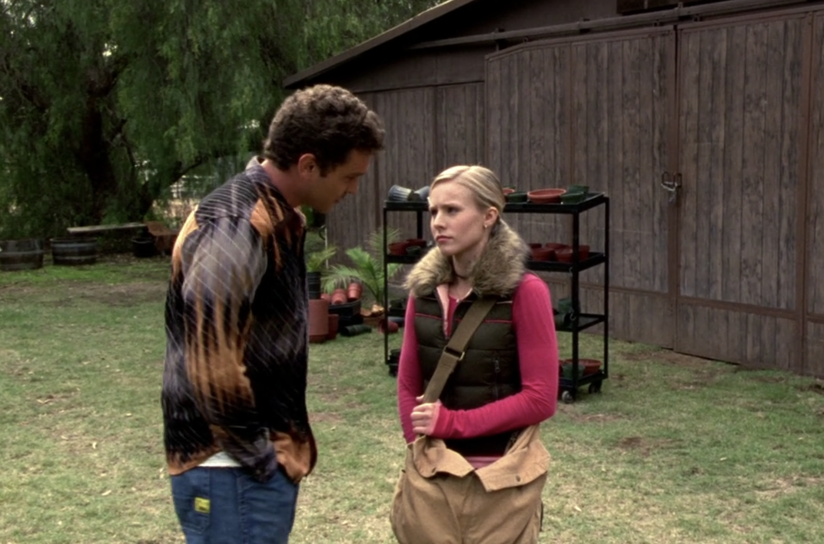 Screenshot from S1E9 of Veronica Mars. Josh and Veronica stand at the farm. Josh is wearing an orange and gray buttondown shirt. He is standing close to Veronica and his head is bent towards her while he explains something. Veronica looks up at him in skepticisim, one hand in her shoulder bag. She his wearing a longsleeved pink shirt and brown vest with a fur collar.