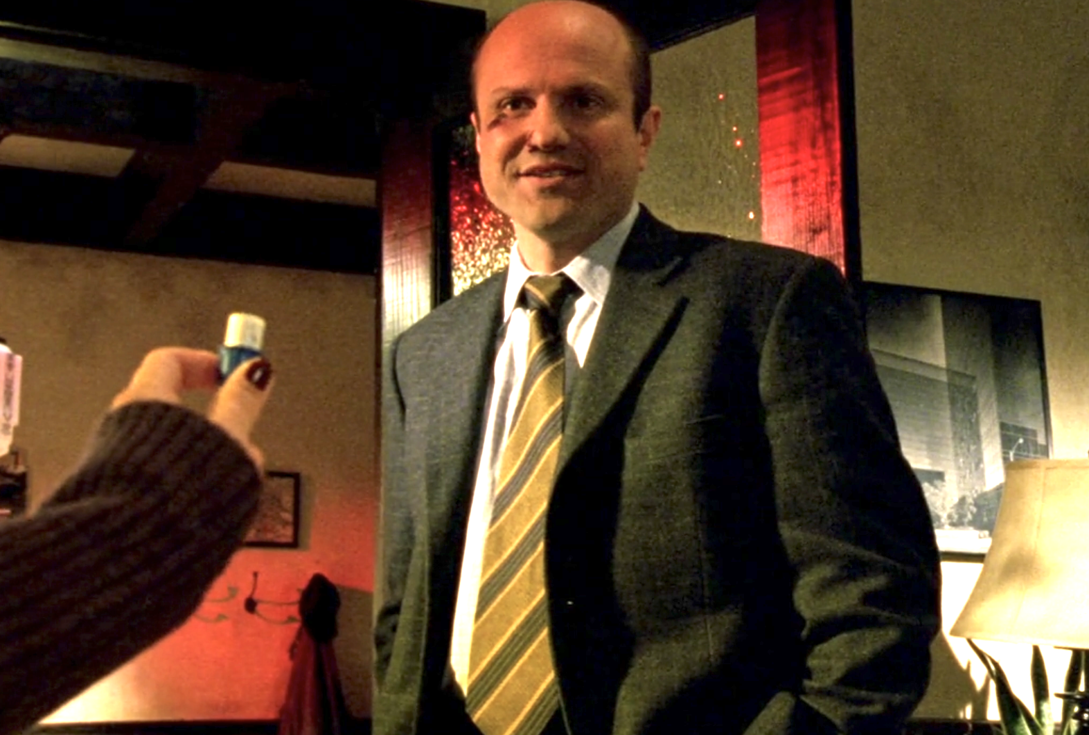 Screenshot from S1E9 of Veronica Mars. Enrico Colantoni as Keith Mars is standing in at Veronica's desk inside Mars Investigations. He is wearing a suit and looking at Veroncia's outstretched hand which is holding a vial. Keith is smiling a little. He has a bruise under his right eye.