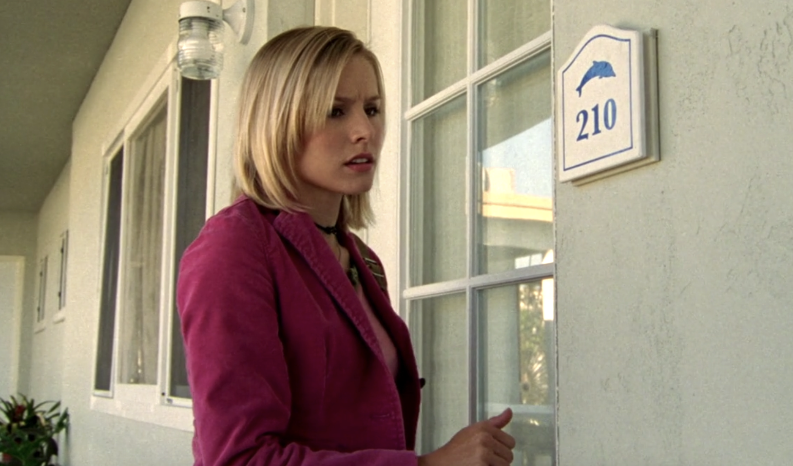 Screenshot of S1E7 of Veroncia Mars. Veronica is wearing a pink blazer and is standing outside the door of an apartment with a nameplate that reads "210" with an image of a dolphin. Her hand is in a fist by the door as if she has just knocked or is about to.
