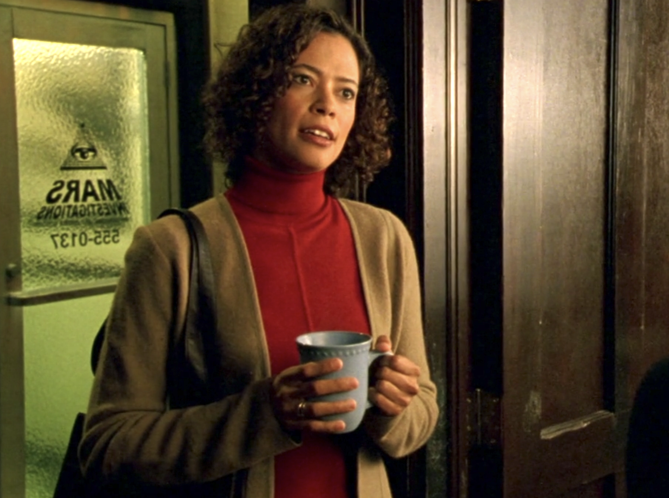 Screenshot from S1E8 of Veroncia Mars. Alicia Fennel is standing in Mars Investigations holding a light blue mug. She's wearing a reddish turtleneck and a brown cardigan.