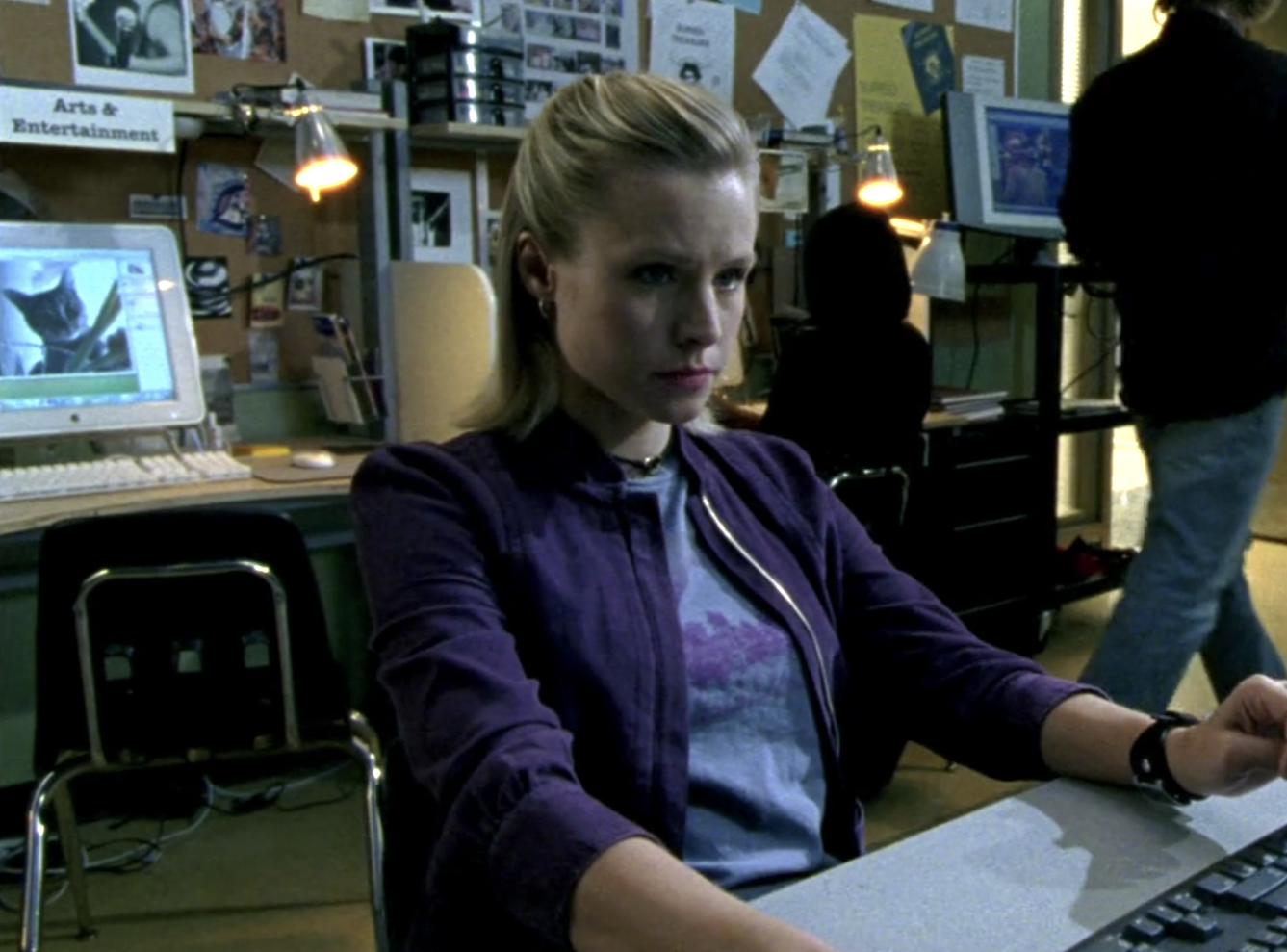 Screenshot from S1E7 of Veronica Mars. Veronica is sitting in a classroom at a computer. She is wearing a purple jacket. On a computer behind her is an imagine of a cat.