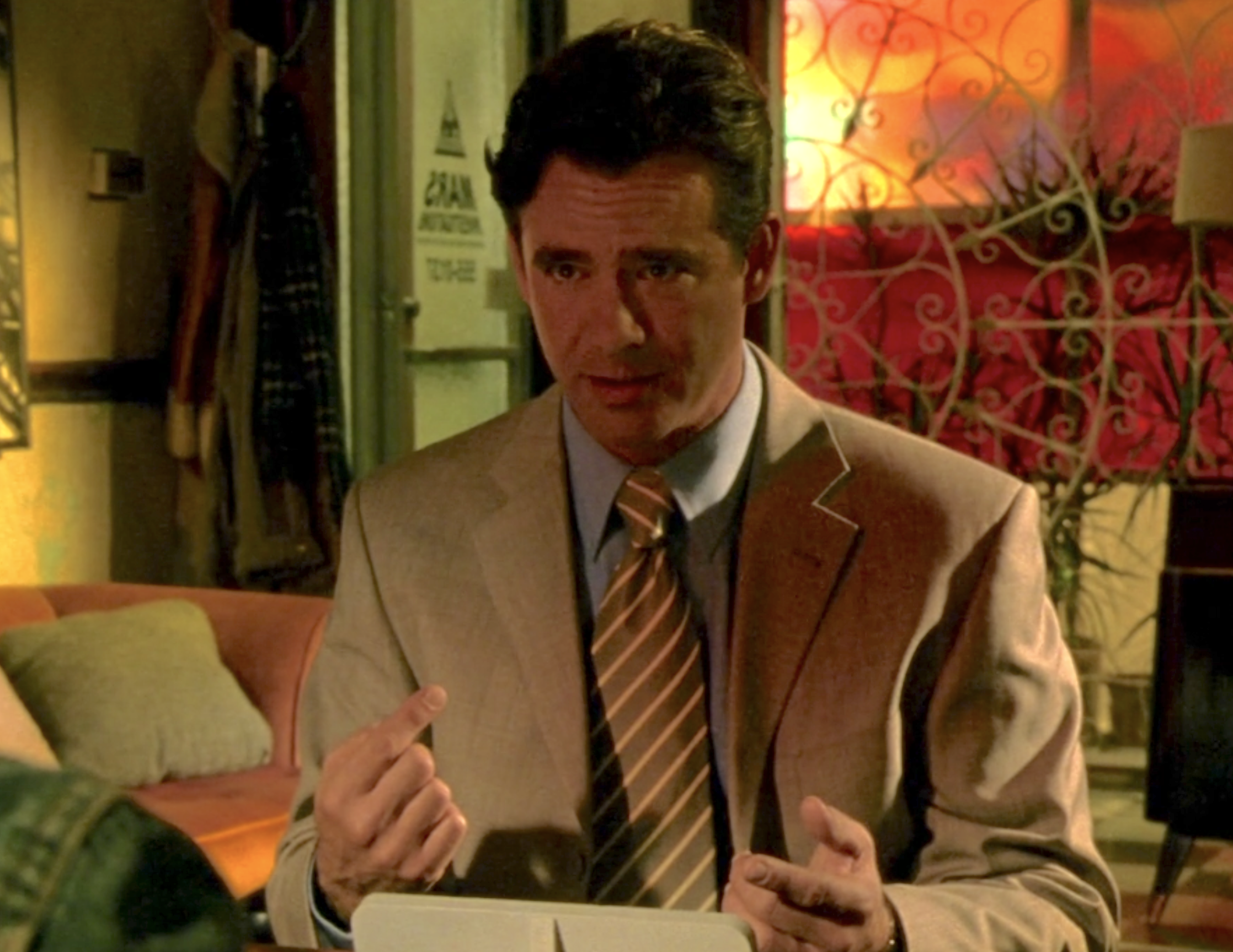 Screenshot from Veronica Mars S1E8. Cliff McCormack is sitting across from an offscreen Veronica in the waiting room of Mars Investigations. He's wearing a tan suit with a blue collared shirt and a tie with browna and white stripes. He is pointing to himself as he talks.