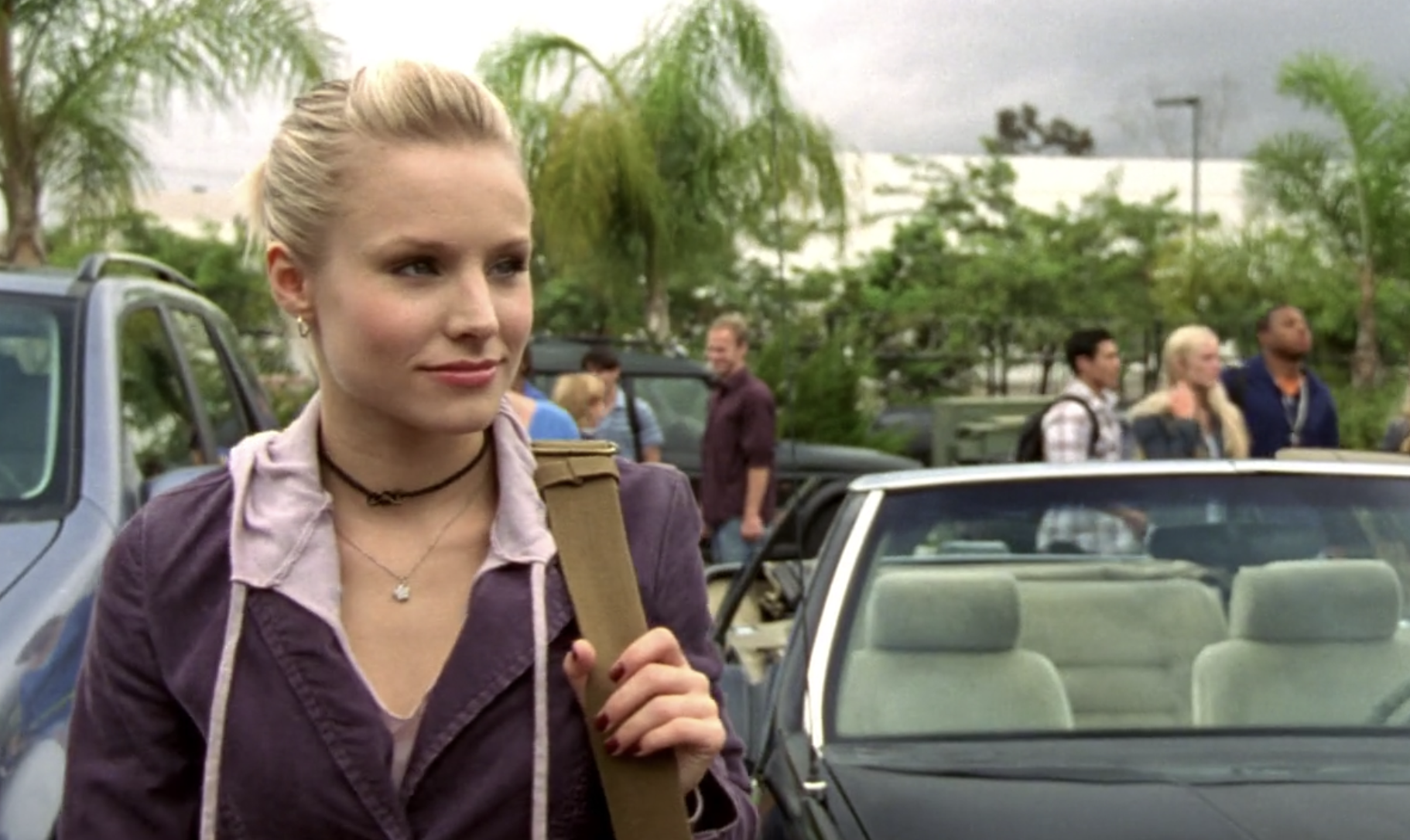 Screenshot from S1E9 of Veronica Mars. Veronica is standing by her car in the Neptune High parking lot. She's wearing a purple jacket and has her taupe shoulder bag over her shoulder. She is looking with a wry smile at an off-screen Duncan.