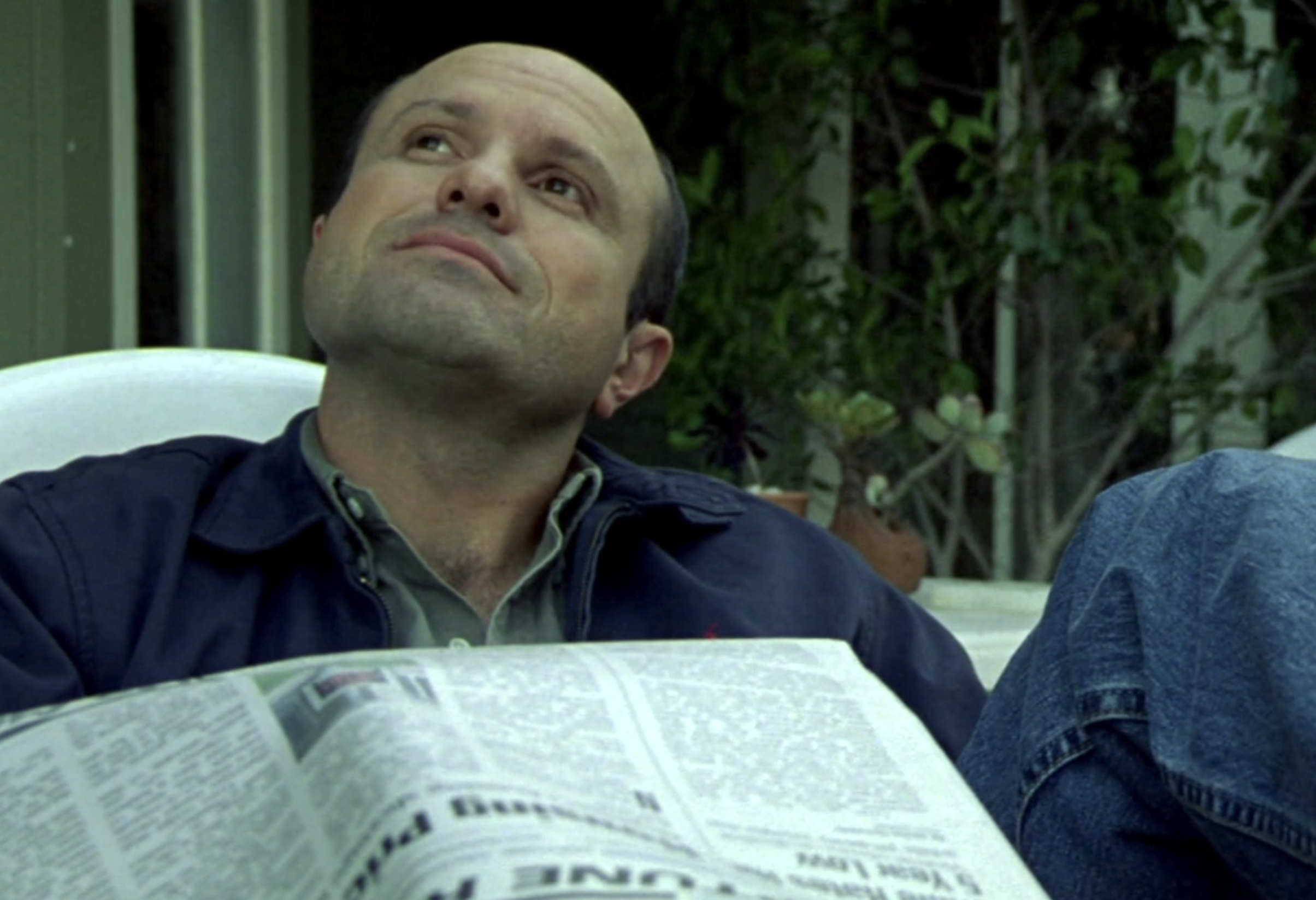 A screenshot from S1E8 of Veronica Mars. Keith Mars is seated outside in a blue jacket and jeans. His head is tilted up and to our left, he's looking at the sky. A newspaper sits on his lap. He has a look of relaxed satisfaction on his face.