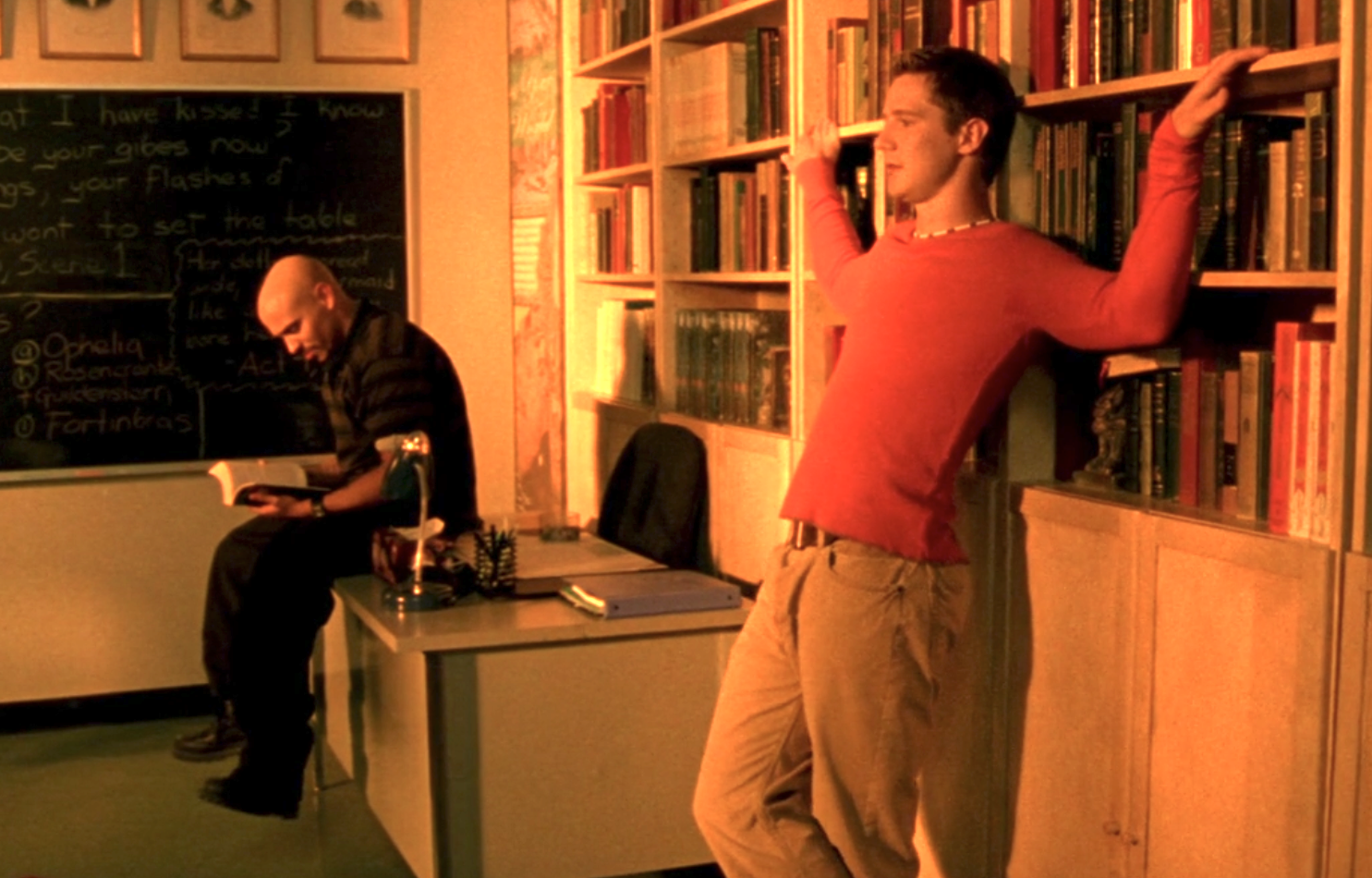 Screenshot from S1E7 of Veronica Mars. Weevil is in the background dressed in black, reading a book while sitting on a desk. Logan is in the foreground in an orange shirt, his arms raised to the sides and leaning on the bookshelf behind him.