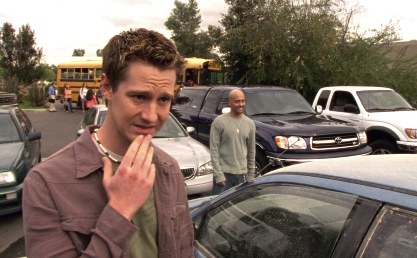 A screenshot from S1E7 of Veronica Mars. Logan is in the foreground in a brown button down shirt with an army green shirt underneath. He is looking at an off-screen character with a look of mock concern, his hand on his chin. Weevil is in the background laughing. They're in a parking lot.