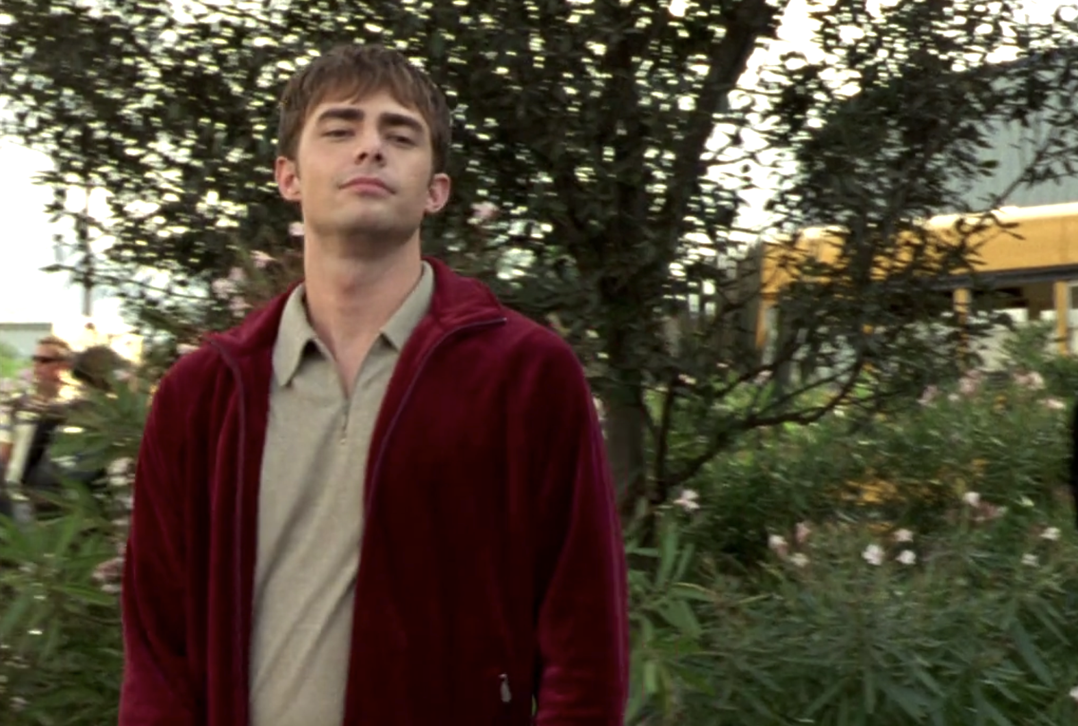 Screenshot from S1E9 of Veronica Mars. Casey Gant in a deep red zip up jacket has his head tilted slightly back and is smirking a little bit, his eyes narrowed. He's in the parking lot at Neptune High.