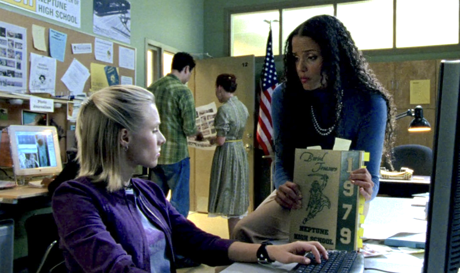 A screenshot of S1E7 of Veronica Mars. Veronica is seated in a classroom at a computer in a purple jacket. She's looking to her side at Ms. Dent who is sitting beside her on the table holding up the 1979 Neptune High School yearbook called "Buried Treasure."