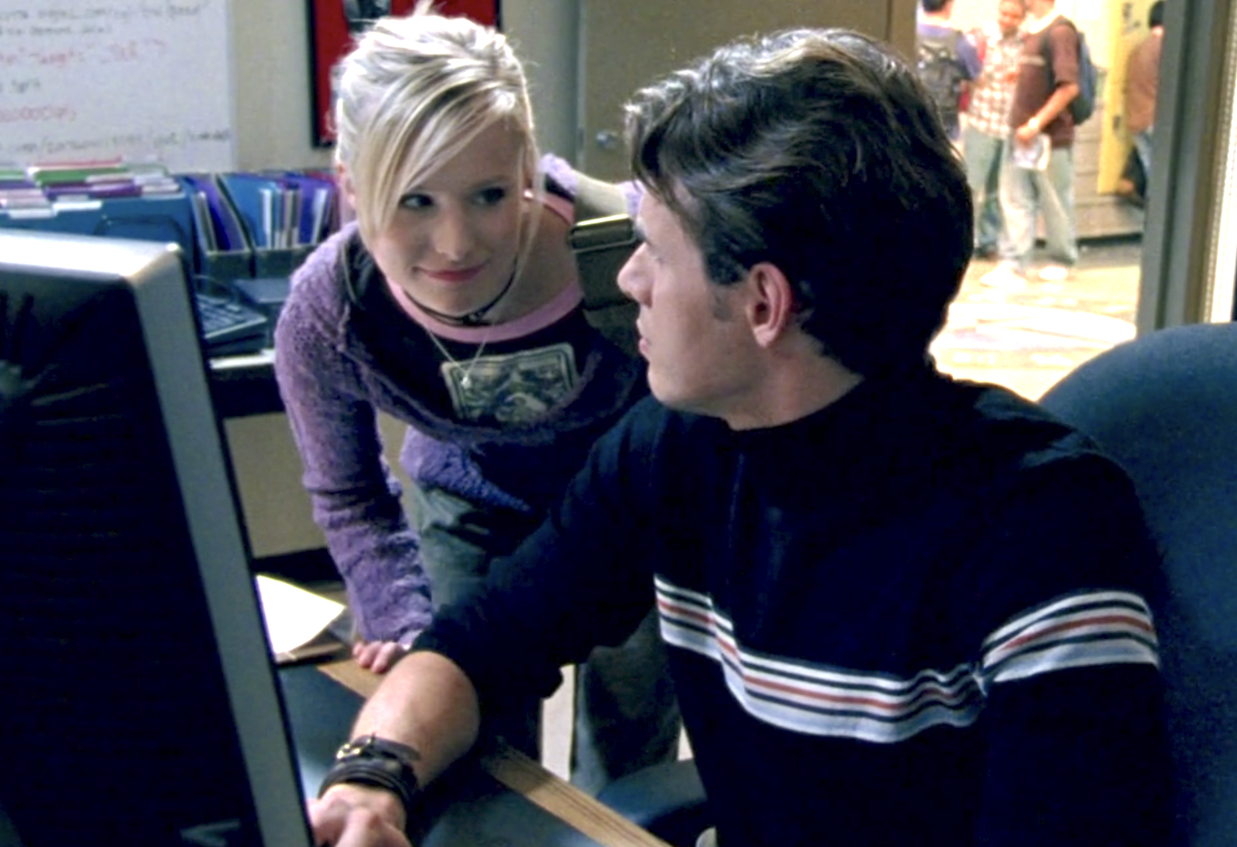 Screenshot from S1E8. Veronica is standing over Renny's shoulder but leaning down and looking at him and smiling. He has turned to his right to look at Veronica. He is seated at a computer.