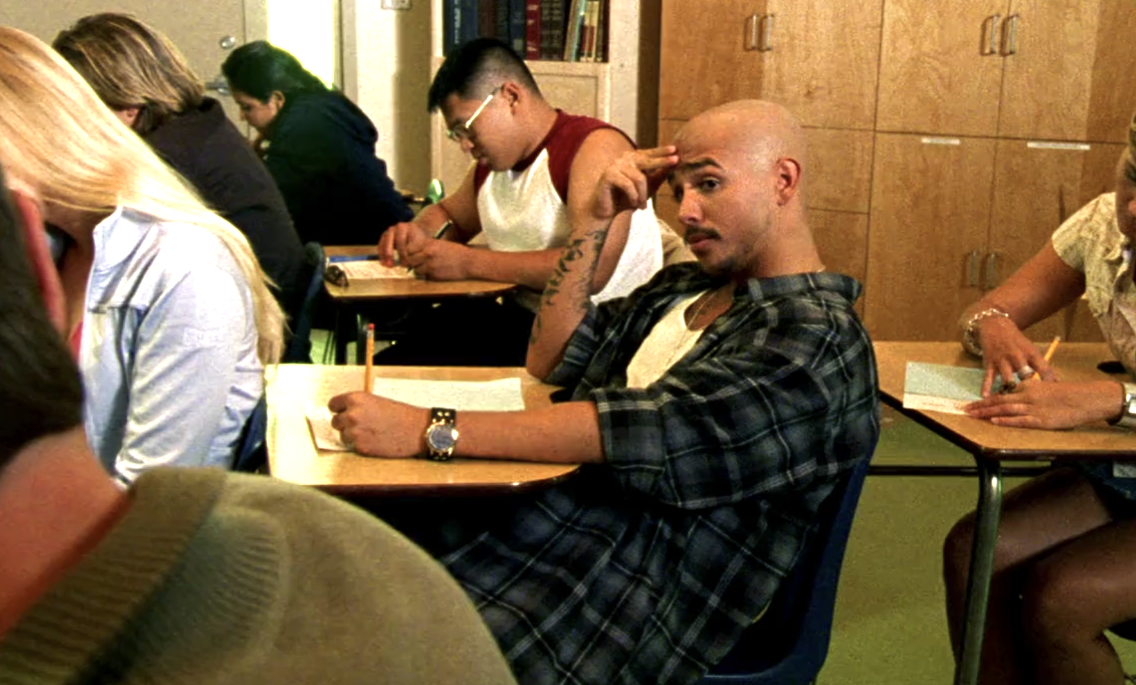 Screenshot from Veronica Mars S1E7. Weevil is sitting in a classroom. He's holding a pencil in his left hand and his hand is resting on a paper on his desk. His right hand is on his forehead and his head is tilted up and he's looking at a desk in the row next to him. He's surrounded by other students taking the test at their own desks.