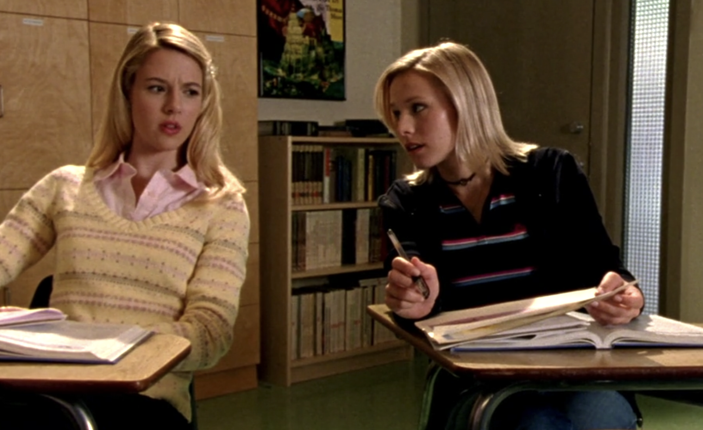 Screenshot of Veronica Mars S1E8. Veronica and Meg are sitting at desks in a classroom. They're leaning towards each other to have a clandestine conversation.