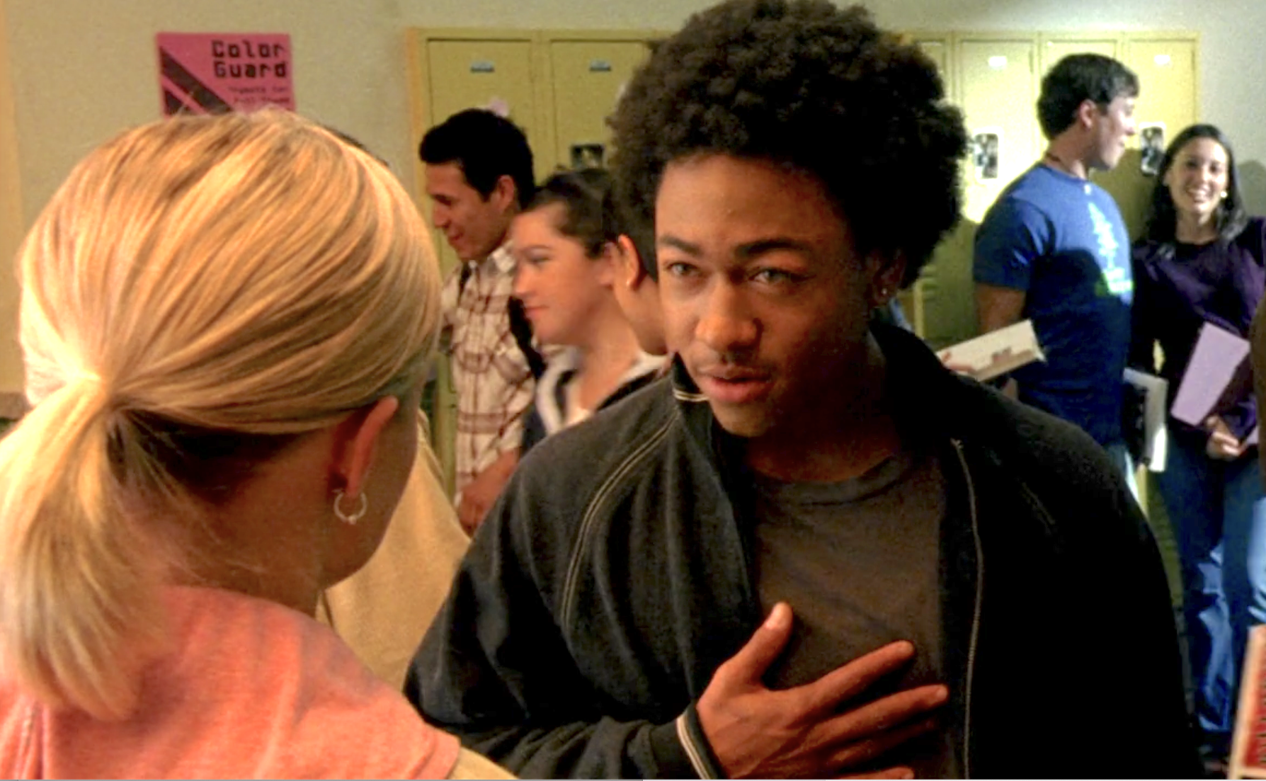 Screenshot from S1E9 of Veronica Mars. Wallace and Veronica are standing in the hallway of Neptune High. Wallace is looking at Veronica. His hand is on his chest and he is making what looks ilke a serious point as Veronica, the back of her head to the viewer, listens.