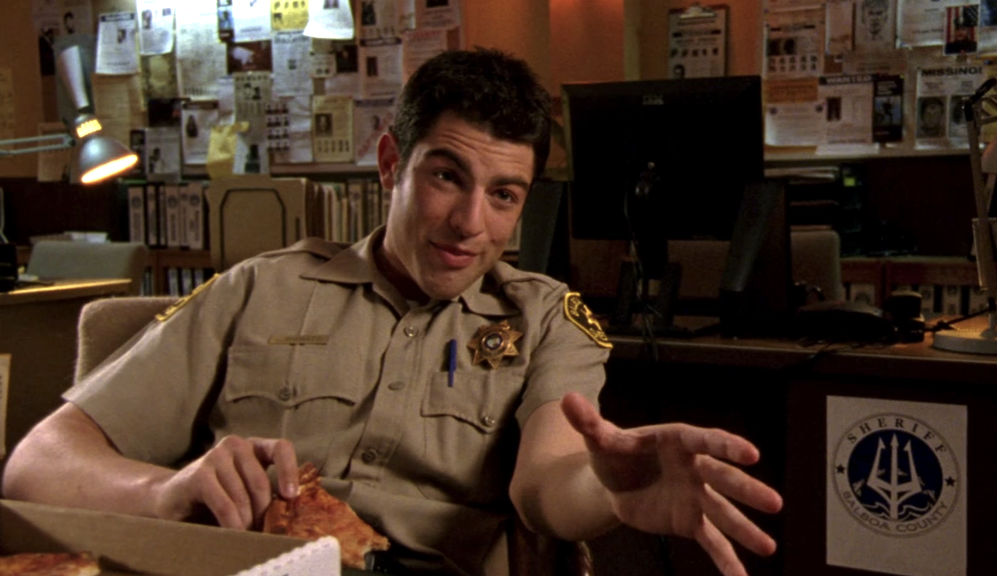 Screenshot of S1E11 of Veronica Mars. Max Greenfield playing Deputy Leo in his beige deputy uniform holds a slice of pizza in one hand and with the other is gesturing at an off-screen Veronica.