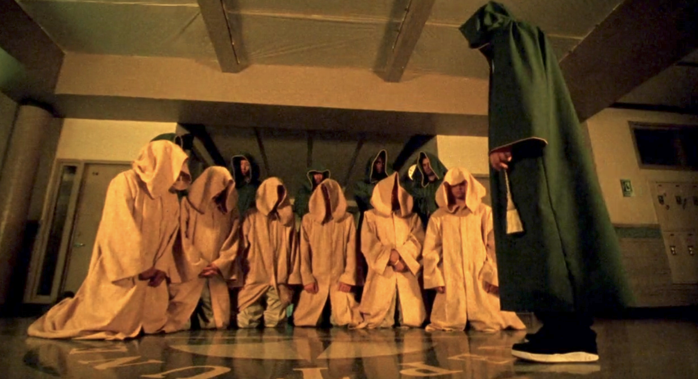 Screenshot from S1E12 of Veronica Mars. Six people in yellow hooded cloaks are kneeling on the floor in a hallway of Neptune High. A figure in a green hooded cloak stands in front and to the side. Five more figures in green hooded cloaks stand behind the kneeling figures.