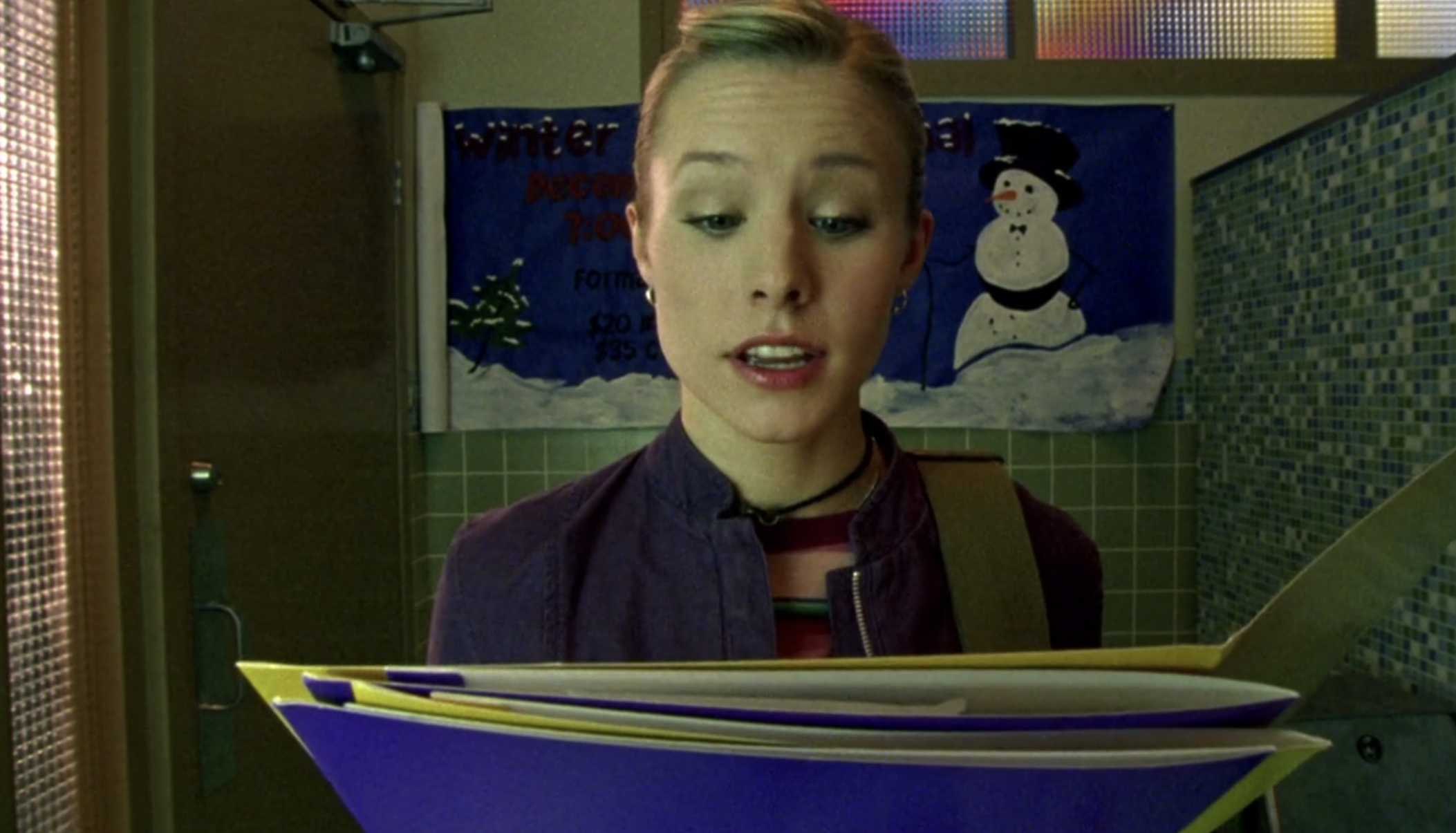 Screenshot of S1E11 of Veronica Mars. Veronica is standing in the bathroom at Neptune High and holding an open folder, looking at it and reading from it.