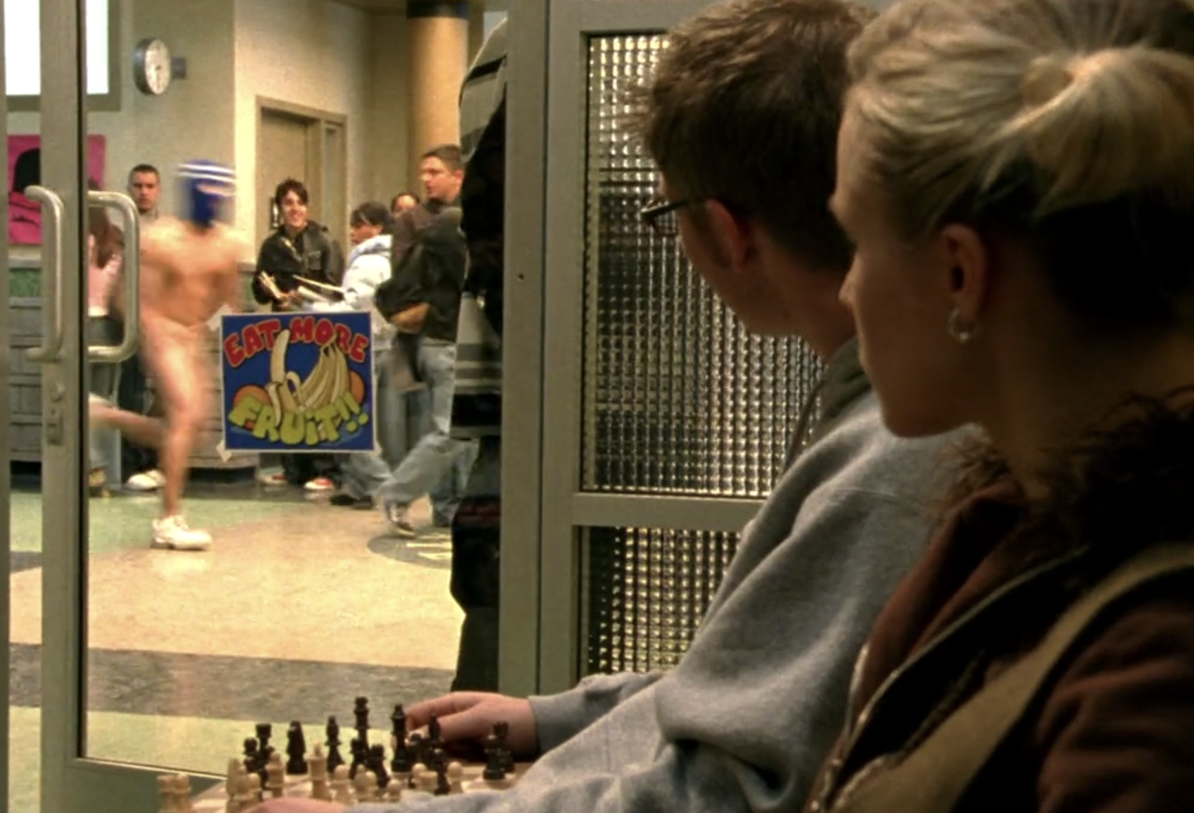 Screenshot from S1E12 of Veronica Mars. Veronica and another student are looking through a glass door at a student running naked down the hallway of Neptune High wearing only a ski mask and sneakers. On the door is a sign that reads "Eat More Fruit!!" with an illustration of bananas and oranges.