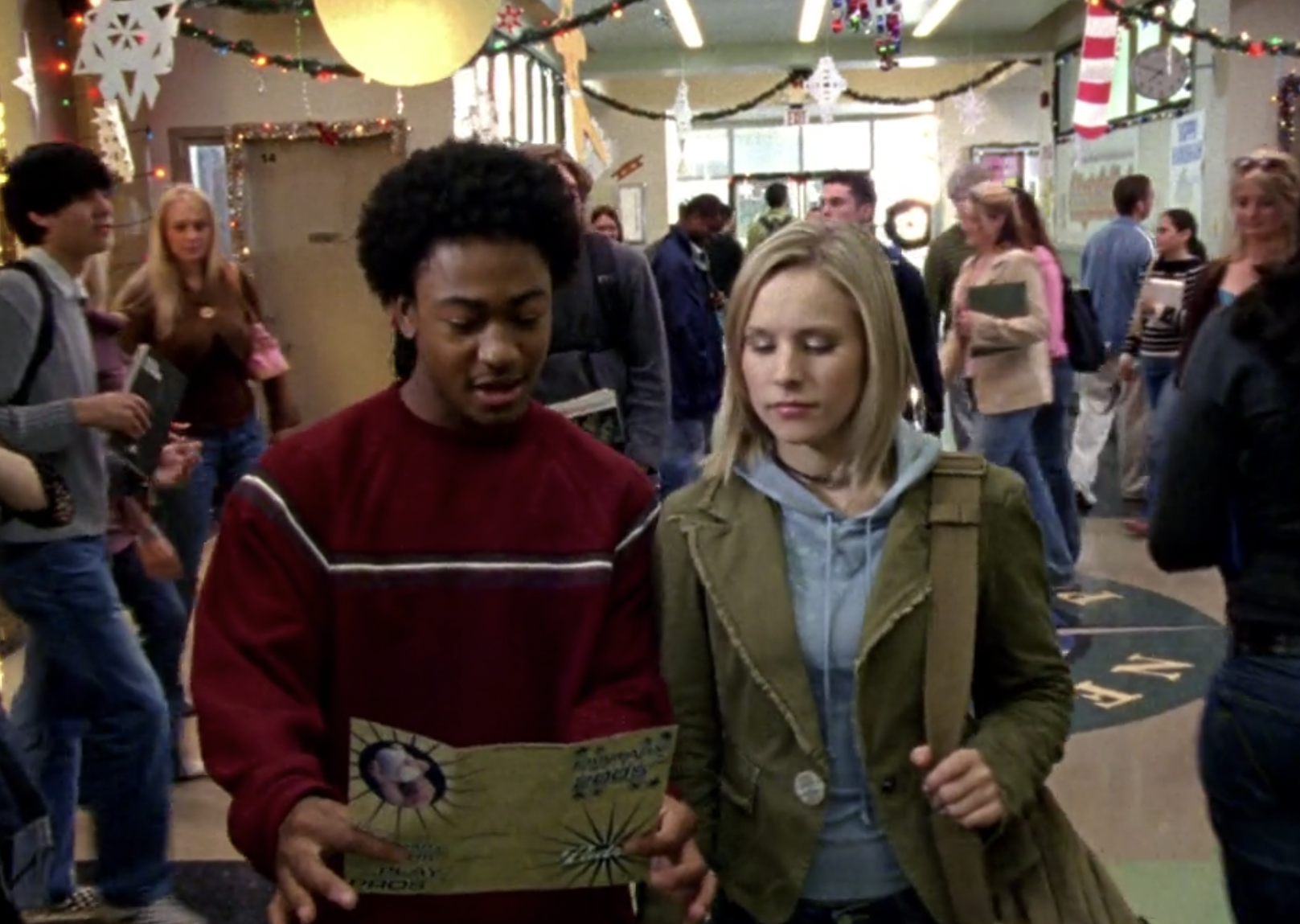 Screenshot from Veronica Mars season one, episode 10. Wallace and Veronica in a crowded hallway at Neptune High looking at a tri-fold pamphlet that Wallace is holding.