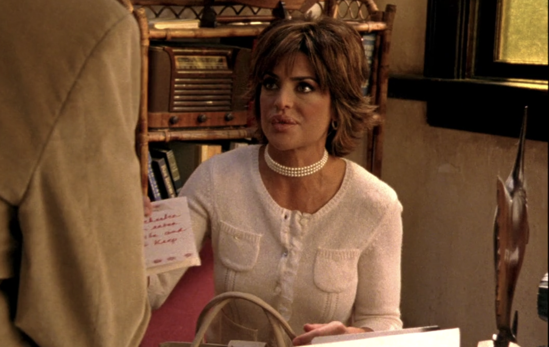Screenshot from Veronica Mars season one, episode 10. Lynn is in Keith's office, wearing a white sweater and a choker made of pearls. She's holding up a threatening note to Aaron written in red marker.