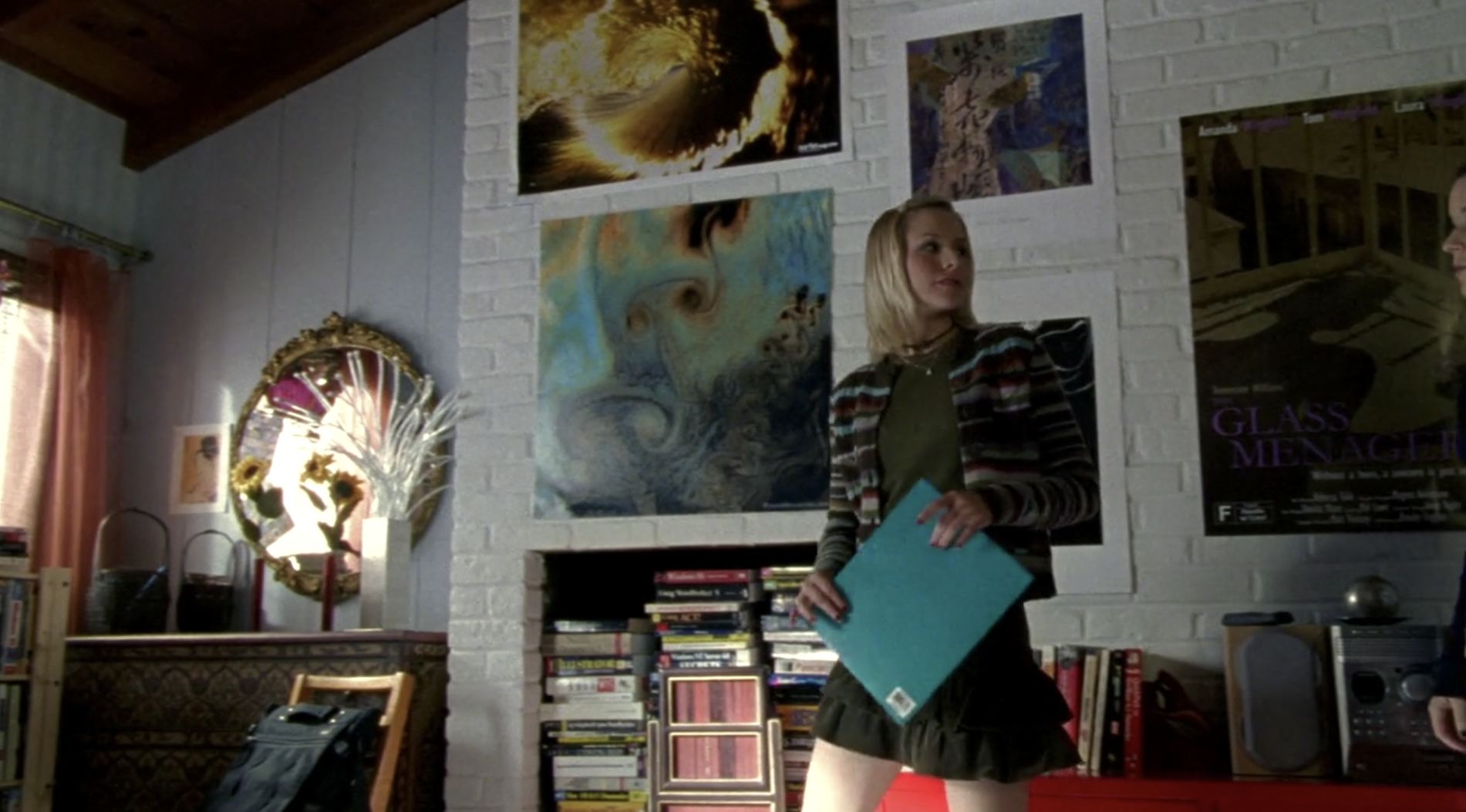 Screenshot of S1E11 of Veronica Mars. Veronica is holding an aqua colored folder and is looking over her shoulder at Mac. They're in Mac's bedroom which has lots of posters on the white brick wall, a circular mirror, some sunflowers, and a lot of books.