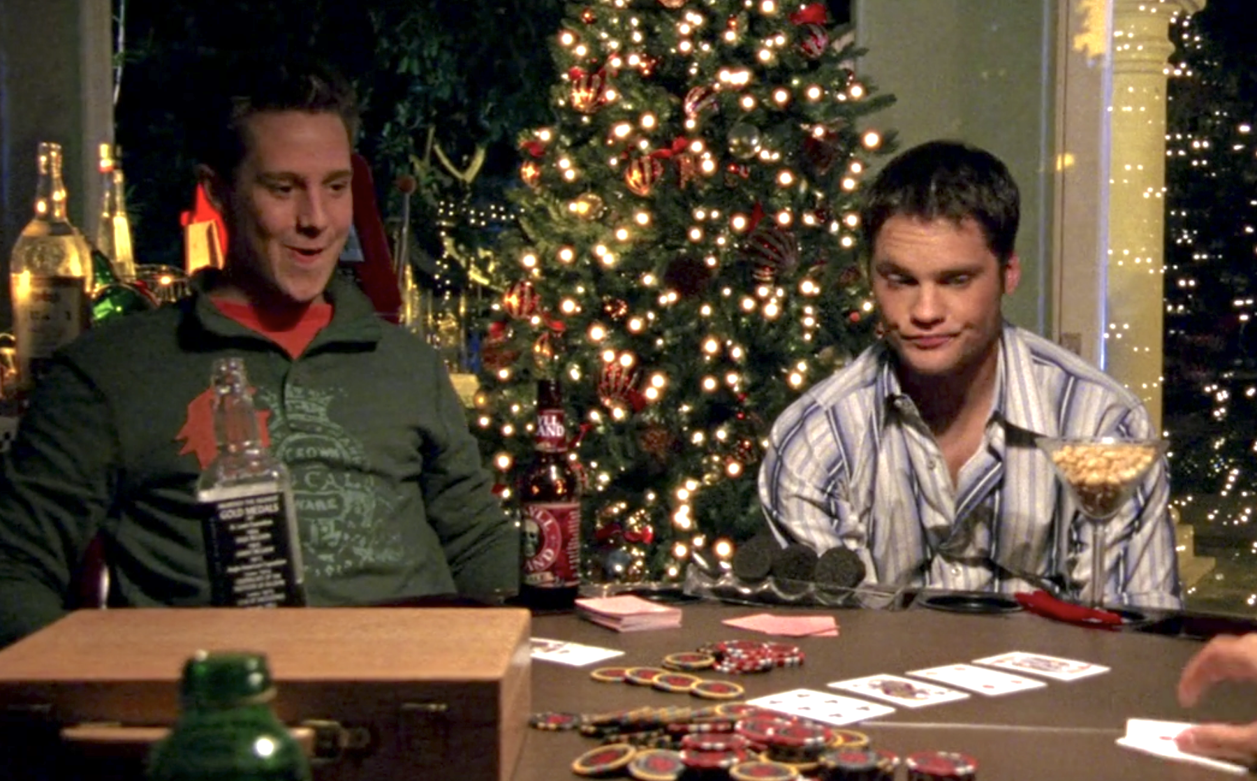 Screenshot from Veronica Mars season one, episode 10. Logan, in a green collared shirt and orange undershirt is laughing and Duncan, in a blue striped collared shirt is looking drunk. They're at a poker table and there's a decorated Christmas tree behind them. The table is spread with poker chips and cards.