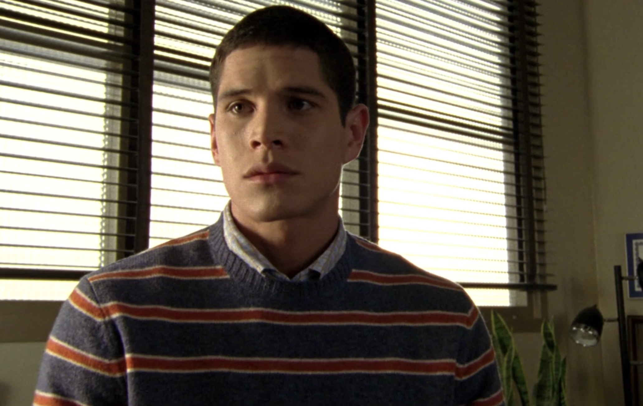 Screenshot of S1E12 of Veronica Mars. Rick is wearing a striped sweater and a collared shirt underneath. He is looking at someone off-screen and looks serious and concerned.
