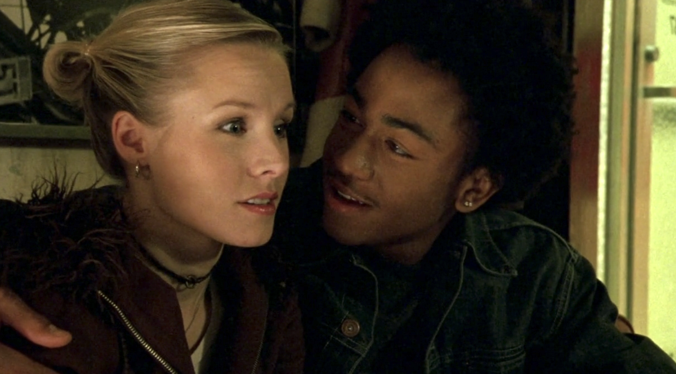 Screenshot from S1E12 of Veronica Mars. Wallace and Veronica are sitting side by side. Wallace has his arm around Veronica and is leaning in sayinng something and smiling. Veronica has a loook of playful shock on her face.