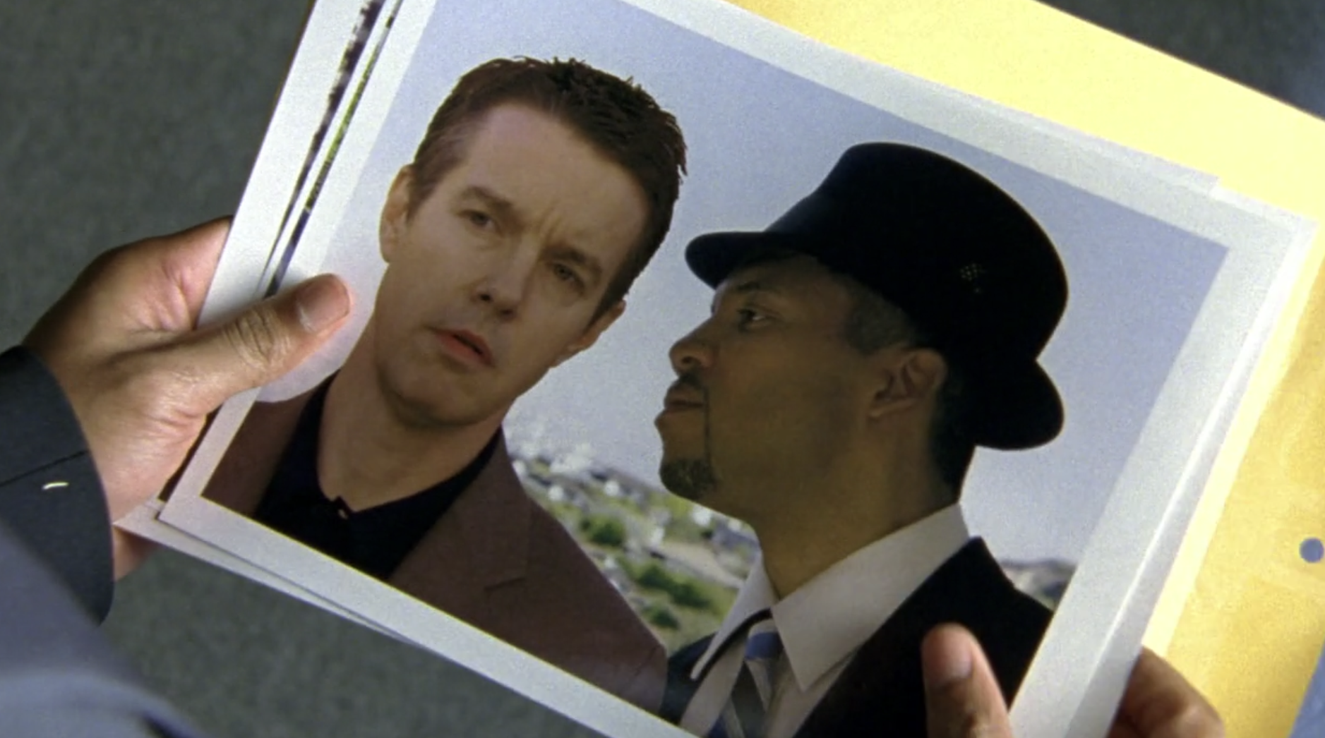 Screenshot of S1E11 of Veronica Mars. Clarence Wiedman's hands holding an 8x10 photograph of Jake Kane staring ahead and Clarence Wiedman beside him in a dark suit and fedora facing sideways and leaning in to say something discreetly to Jake.