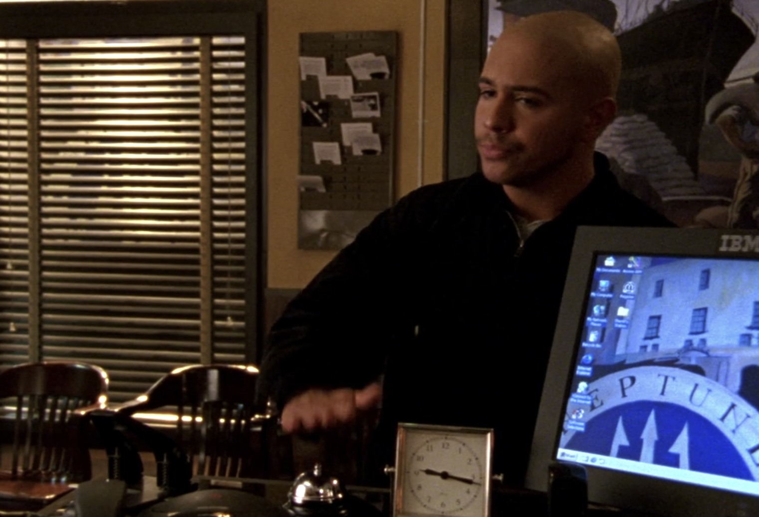 Screenshot of S1E11 of Veronica Mars. Weevil is standing at a desk in the sheriff's department with his hand over a bell he's ringing for service