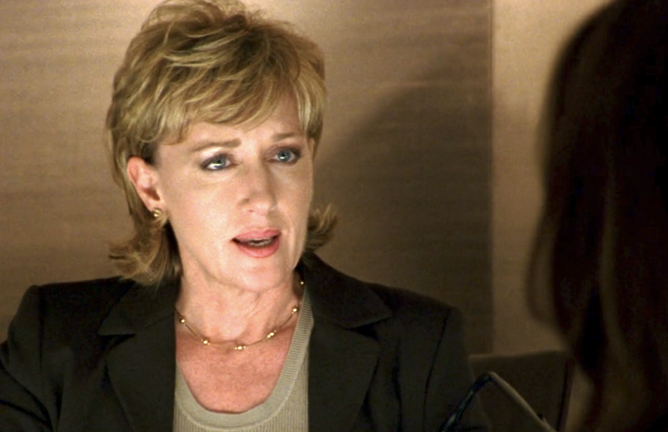 Screenshot from S1E14 of Veronica Mars. A middle-aged woman with short blond hair wearing a gold necklace and earrings and a blazer.