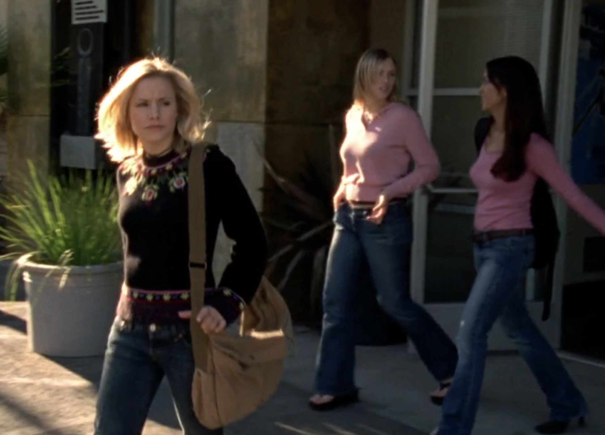 Screenshot from S1E14 of Veronica Mars. Veronica is in the foreground in a brown sweater and carrying a beige shoulder bag. In the background are two young women dressed almost identically in longsleeved, v-neck pink shirts and dark, flared jeans.