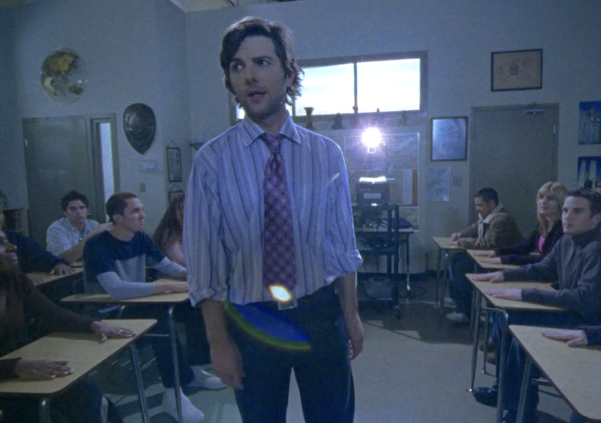 Screenshot from S1E14 of Veronica Mars. Adam Scott in a baggy button down shirt, tie, and baggy jeans with floppy hair is standing at the front of a classroom full of students.