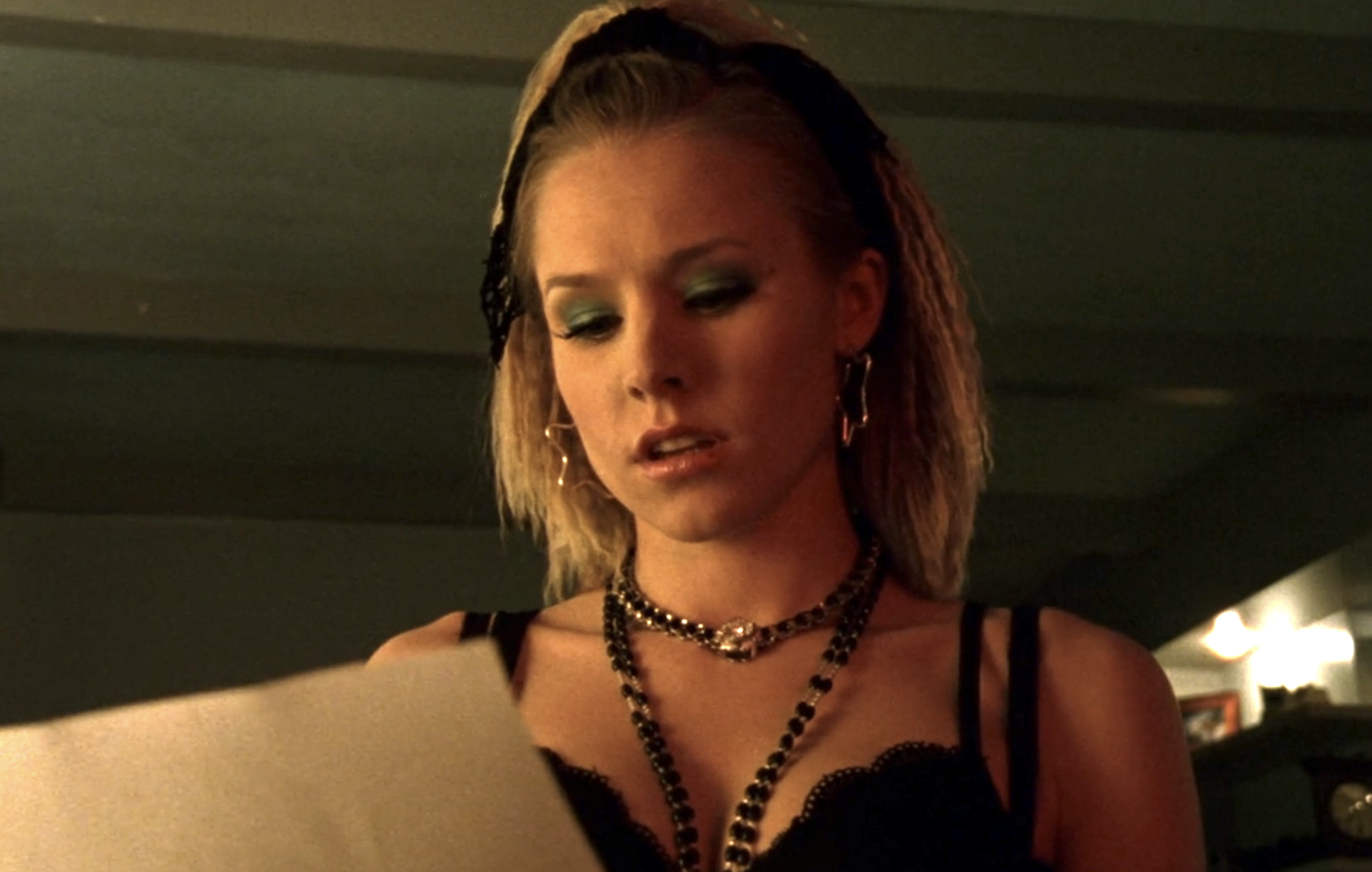 Screenshot from Veronica Mars S1E15. Veronica is dressed like Lucky Star era Madonna. She has crimped hair, black necklaces, big siver earrings, and green eyeshadow. She is looking at a letter.