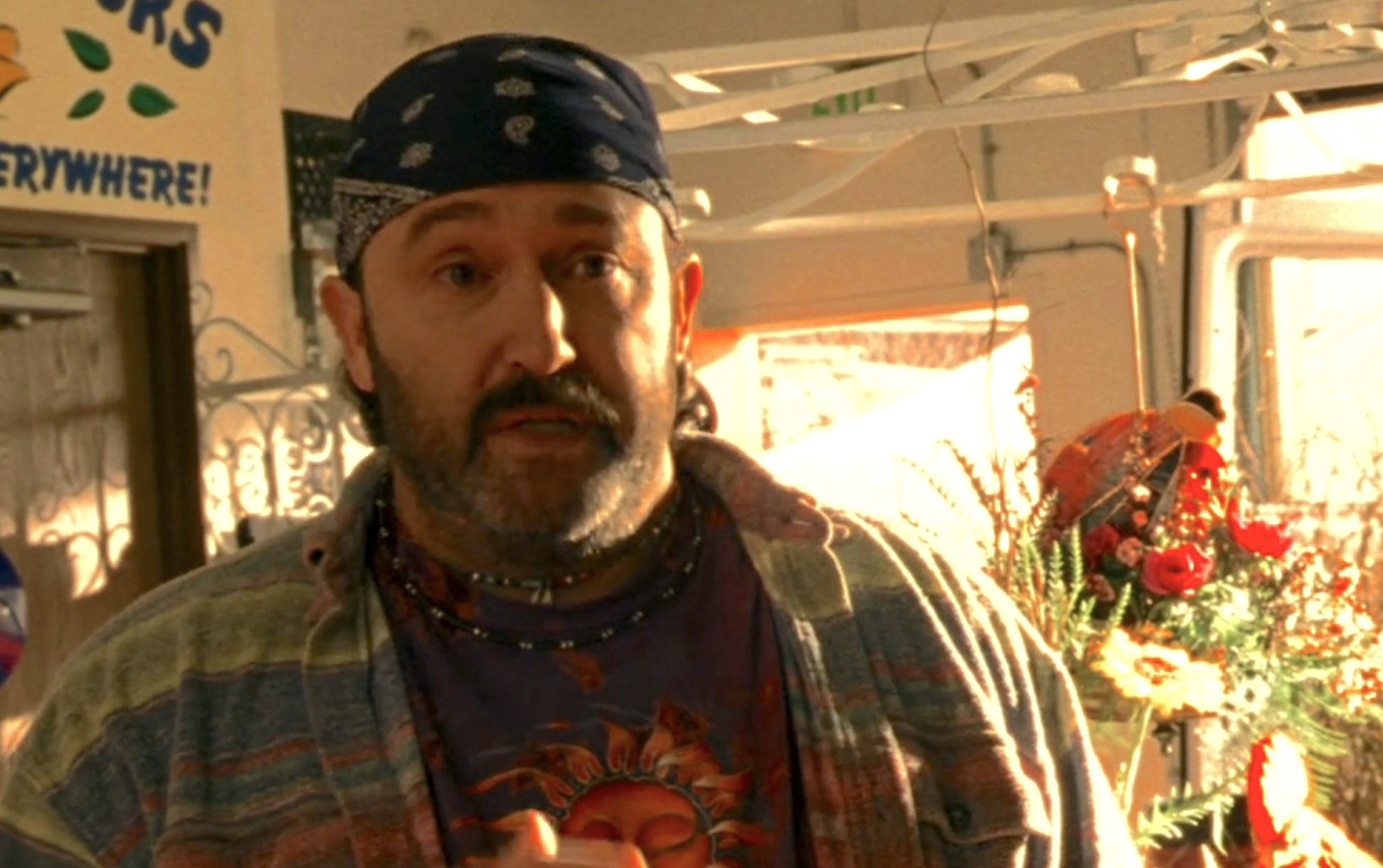 Screenshot from Veronica Mars S1E15. Manny, a man with a bandana, mustache, stubble, flannel shirt is standing in his flower shop talking to Veronica who is off screen.