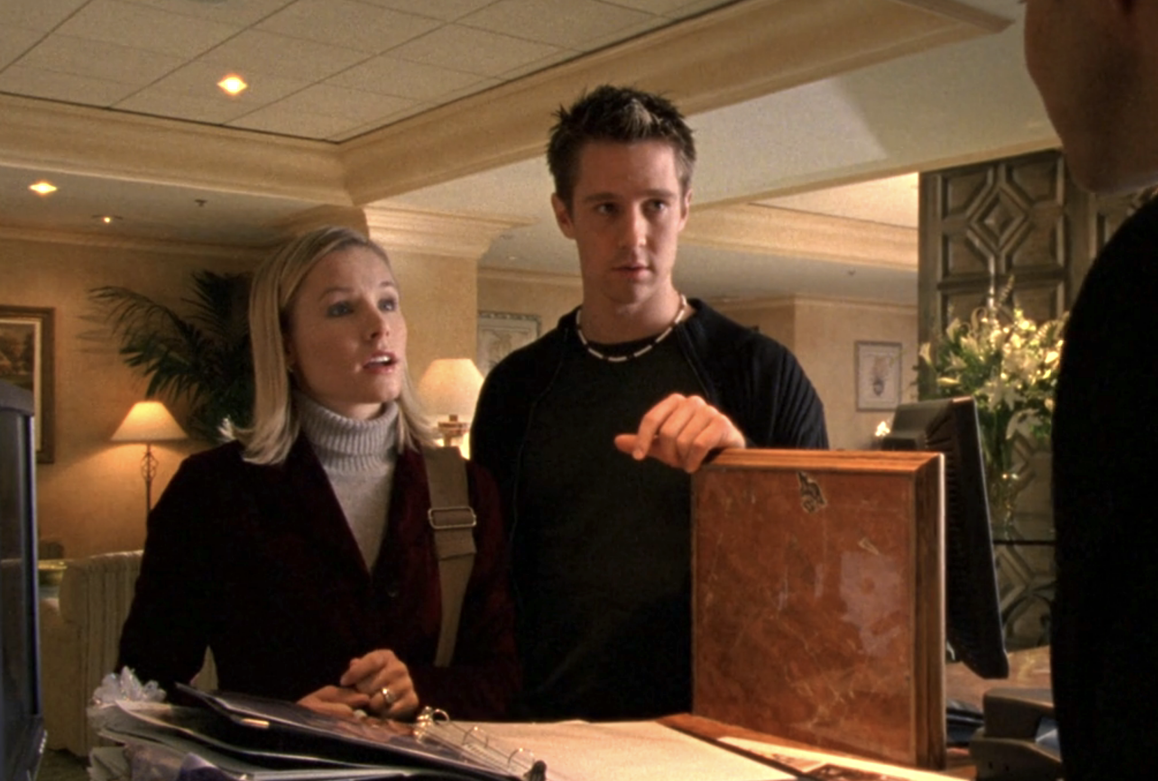 Screenshot from Veronica Mars S1E15. Veronica and Logan are standing at a hotel reception desk. 