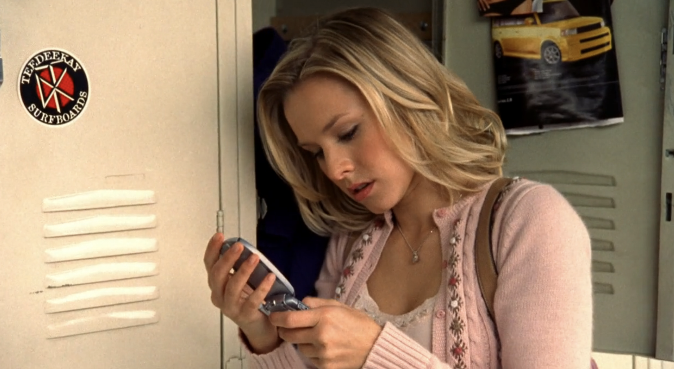 Screenshot from S1E16 of Veronica Mars. Veronica is wearing pink and standing at an open locker looking at a flip phone. To her left there's a sticker on a locker with the Dead Kennedys' logo. It says "Teedeekay Surfboards."