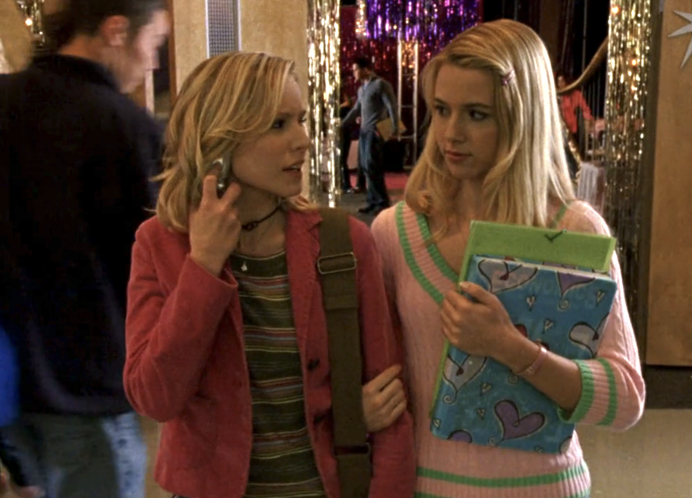 Screenshot from Veronica Mars S1E15 Veronica is holding a flip phone to ear and looking at Meg who is standing beside her, with her hand hooked around Veronica's arm.