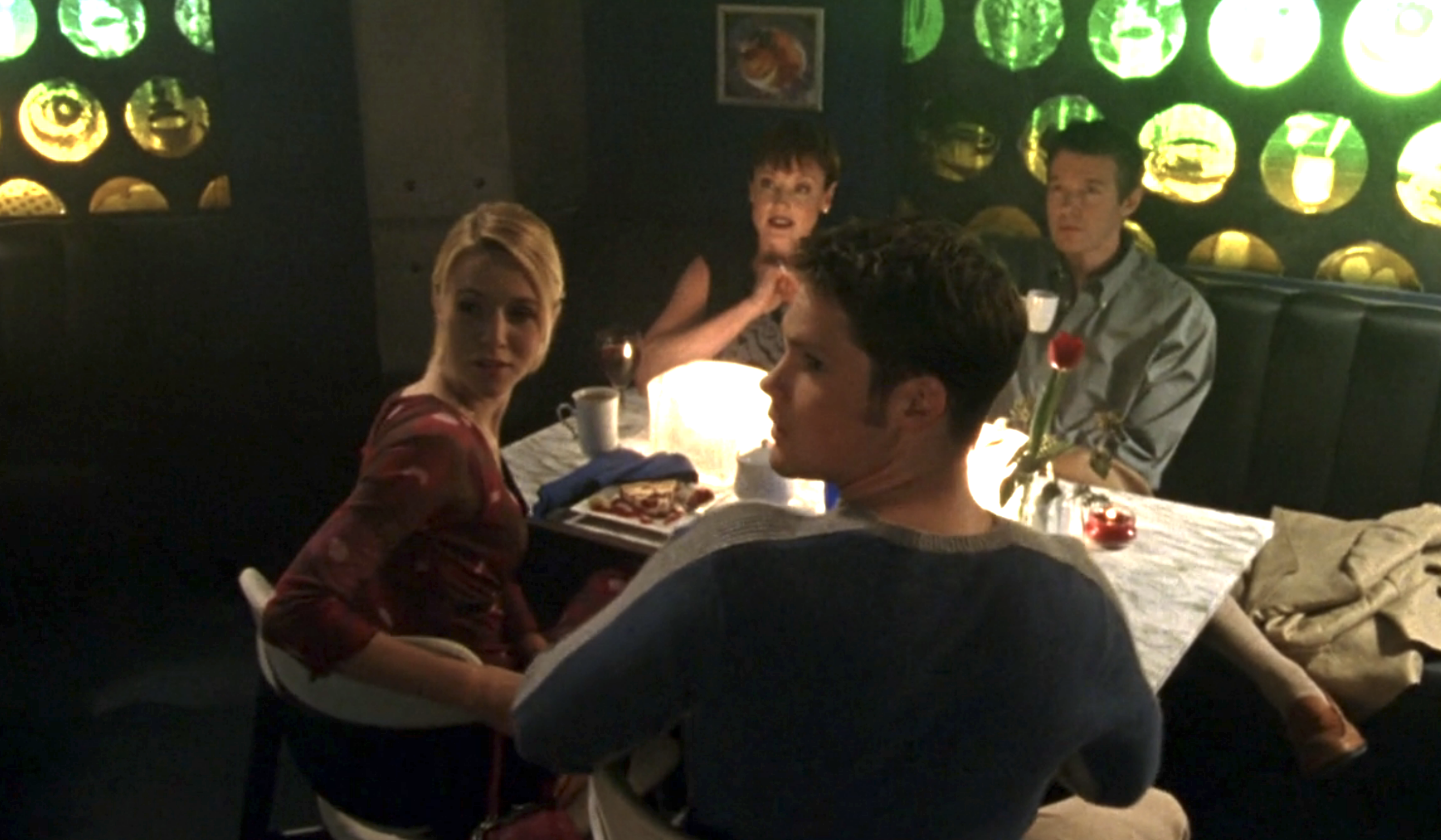 Screenshot from S1E16 of Veronica Mars. Meg, Duncan, and Duncan's parents are sitting at a table in a dimly lit restaruant. They're all looking at an offscreen Veronica and Leo.