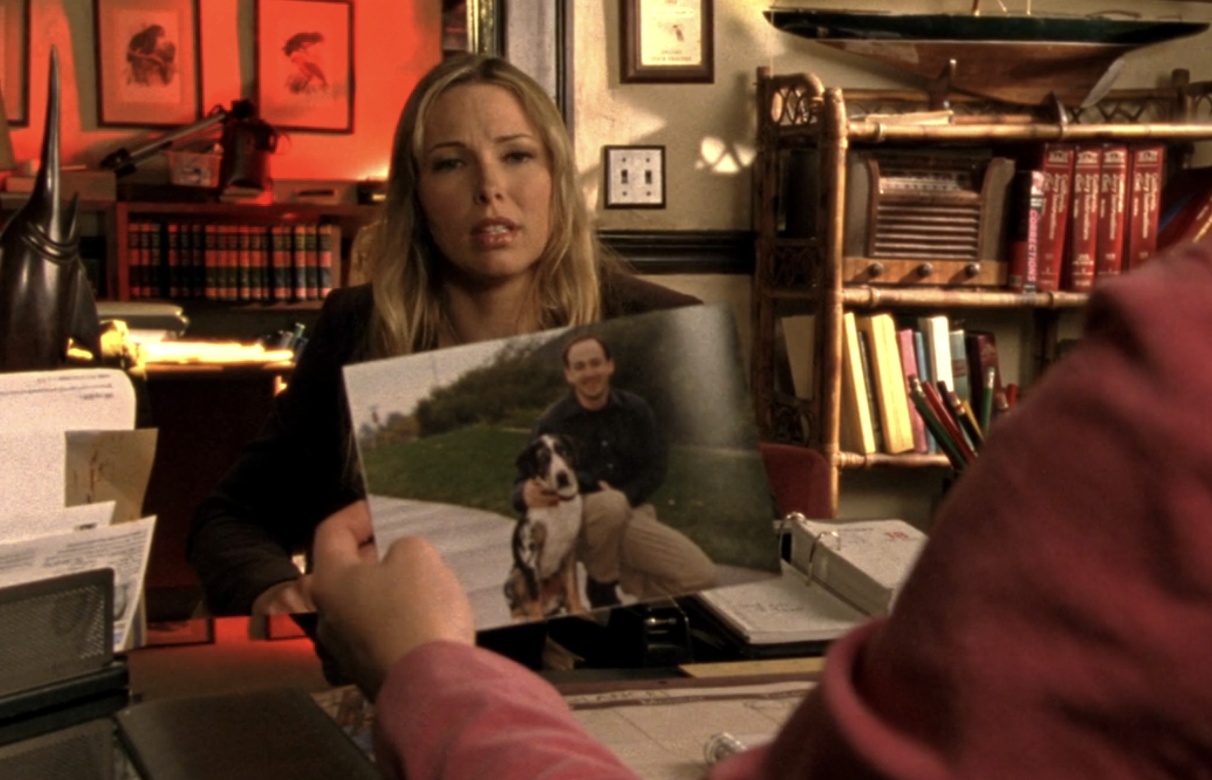 Screenshot from Veronica Mars S1E15. A blond woman is sitting at a desk looking at Veronica who we see from behind holding a photograph of a balding man with a dog.