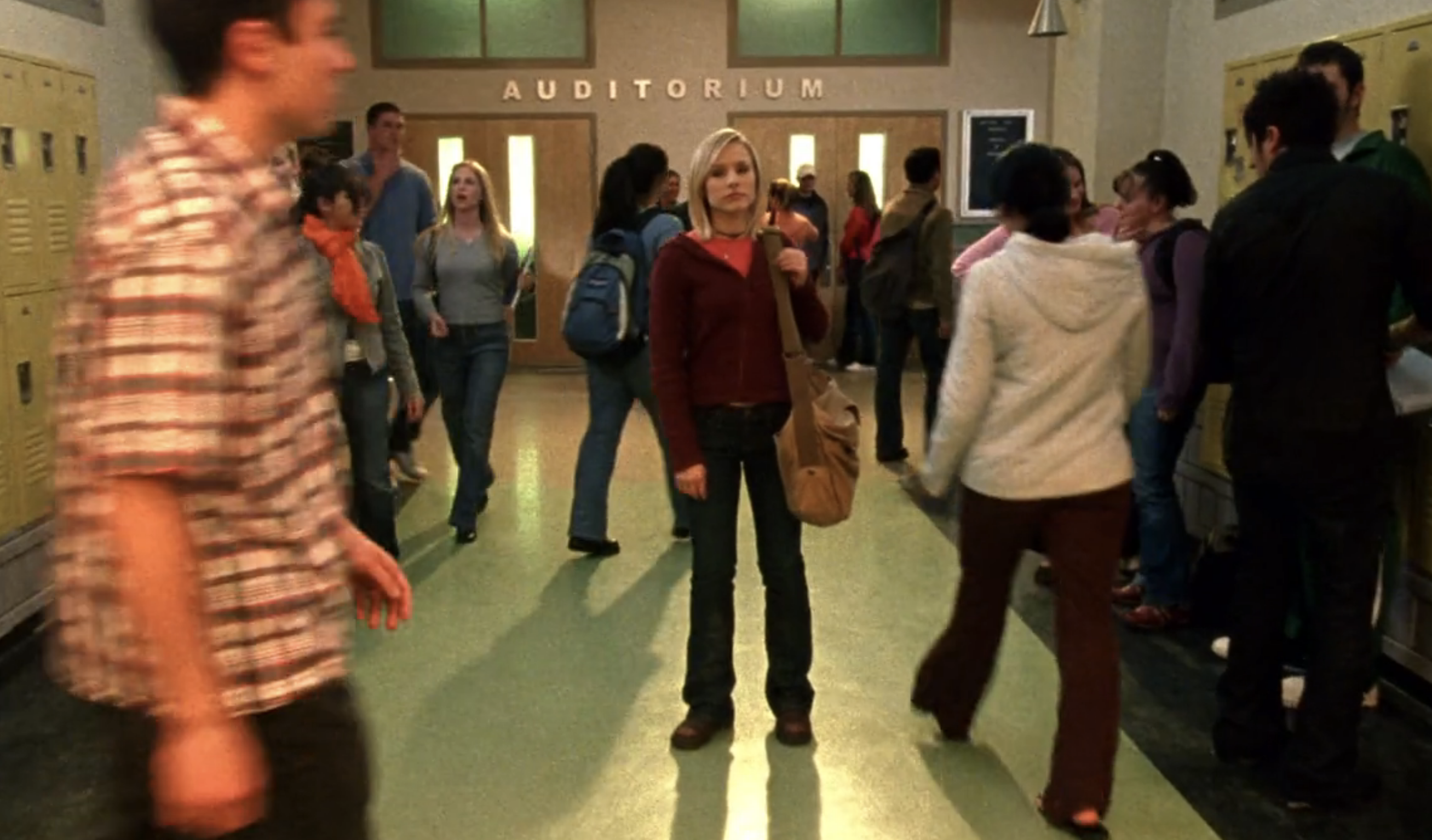 Screenshot from S1E16 of Veronica Mars. Veronica is standing in the busy hallway of Neptune high looking forlornly into the distance