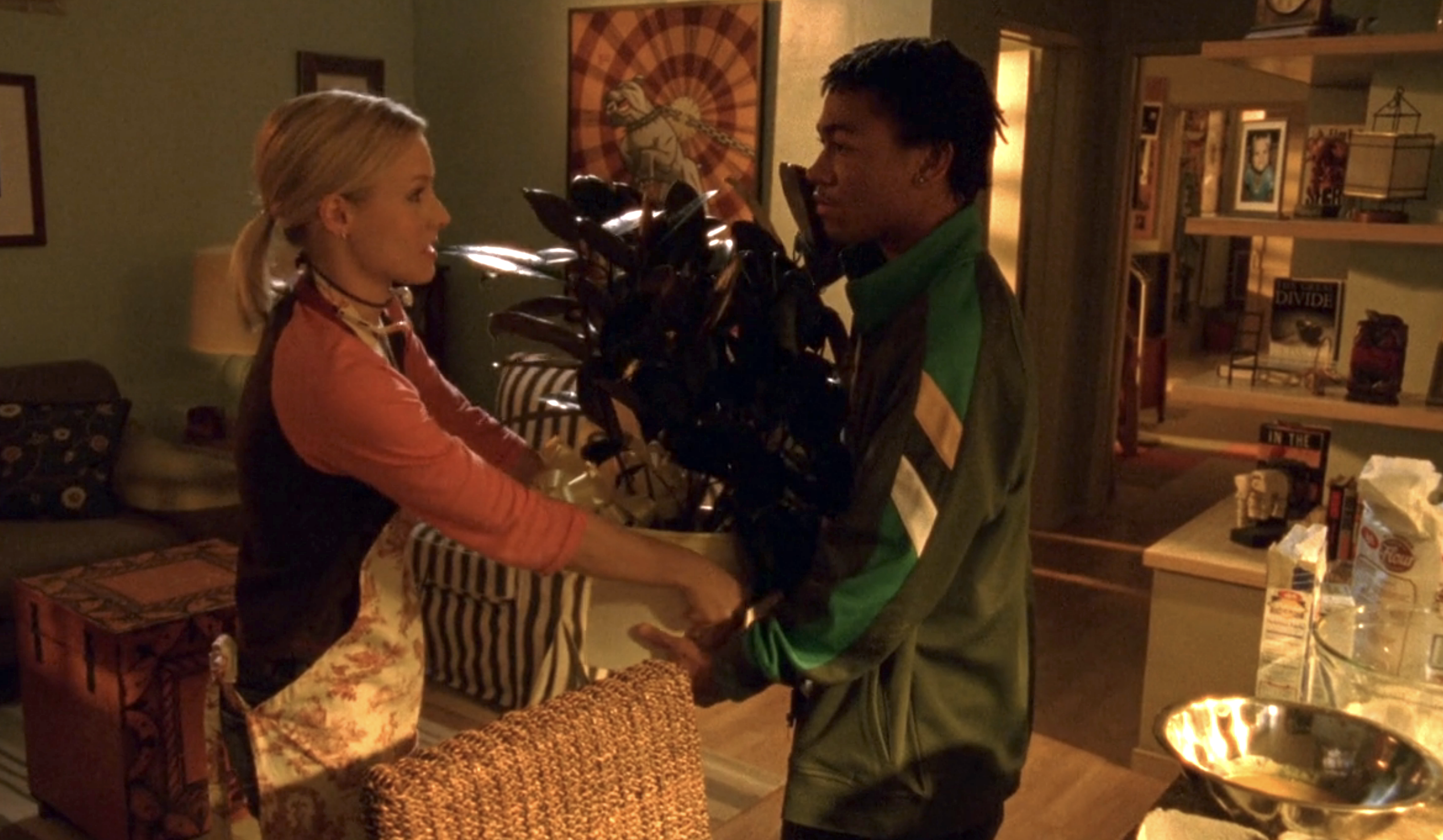 Screenshot from S1E16 of Veronica Mars. Veronica is handing Wallaca a big potten houseplant. They're in her house and she is wearing an apron. He is wearing his green and yellow Neptune High jacket.