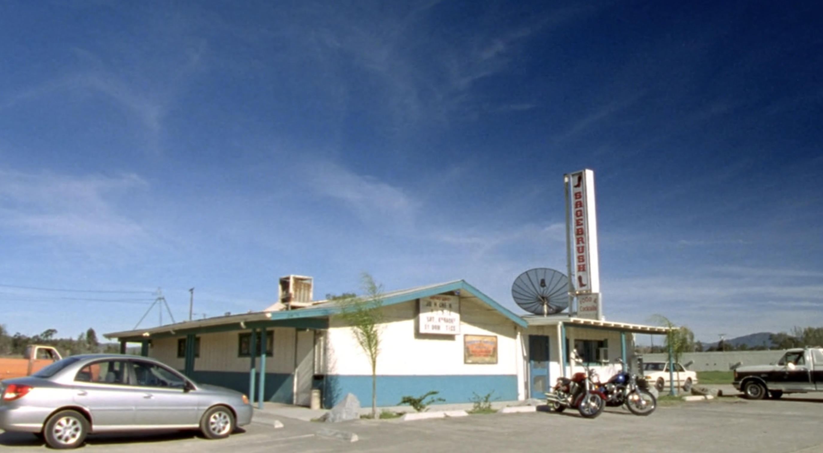 Screenshot from Veronica Mars S1E15. The Sagebrush Cantina, a low white building with blue trim. Two motorcycles are parked outside.